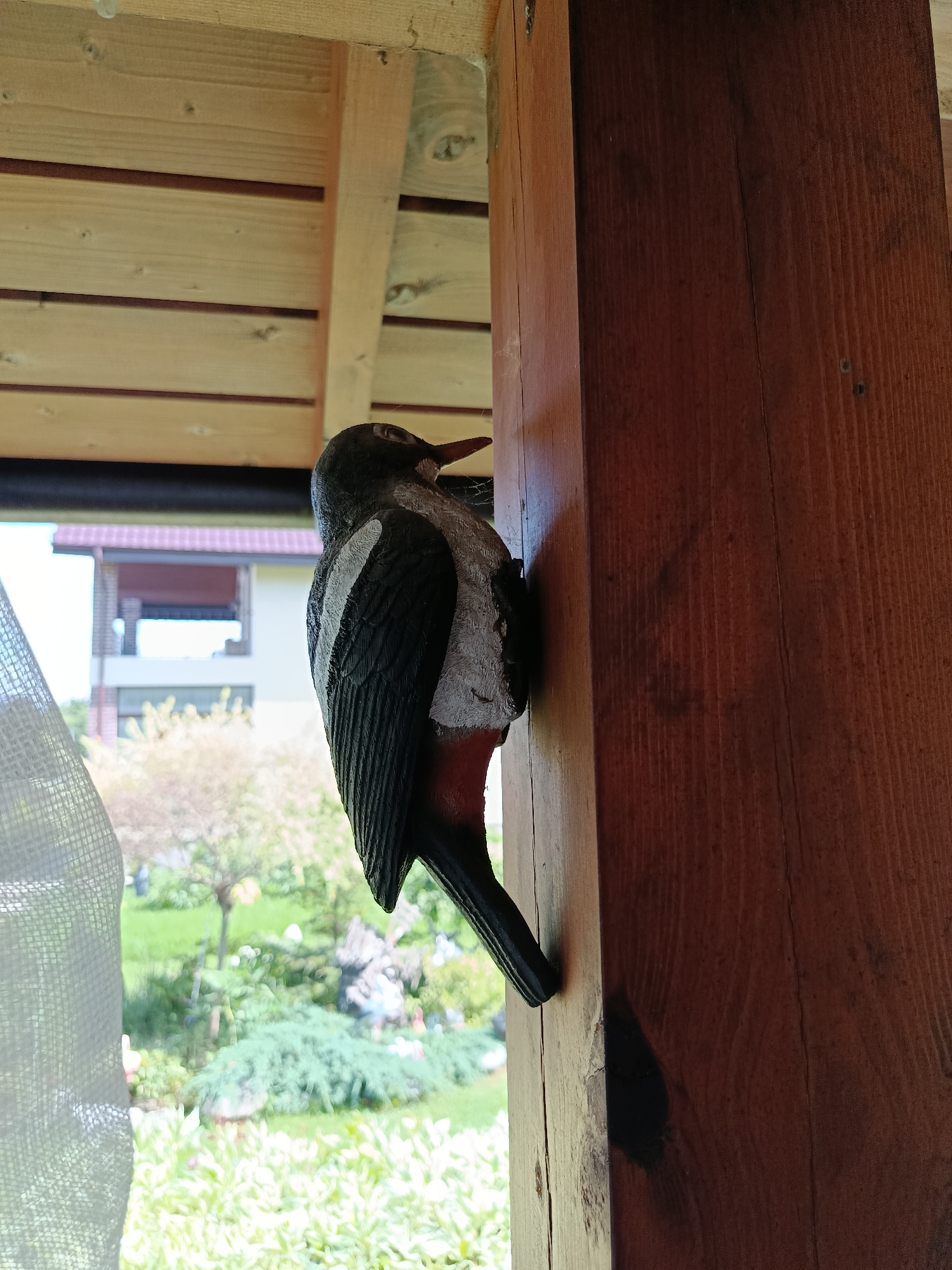 A close-up, eye-level, outdoor shot shows a decorative wooden bird, possibly a woodpecker, mounted vertically on a thick, dark brown wooden pillar. The bird is facing right, with its body angled slightly downwards, and its black and white wings are folded against its back. Its beak is pointed, and its eye is visible. The pillar is part of an overhead wooden structure, with horizontal wooden planks visible in the upper portion of the image. To the left of the pillar, a white, translucent mesh fabric hangs down, partially obscuring the background. The background reveals a blurred garden scene with green grass, bushes, trees, and a distant building with a red-tiled roof. The image appears to be taken on a bright, sunny day. The location is identified as Misaylovo, Russia.