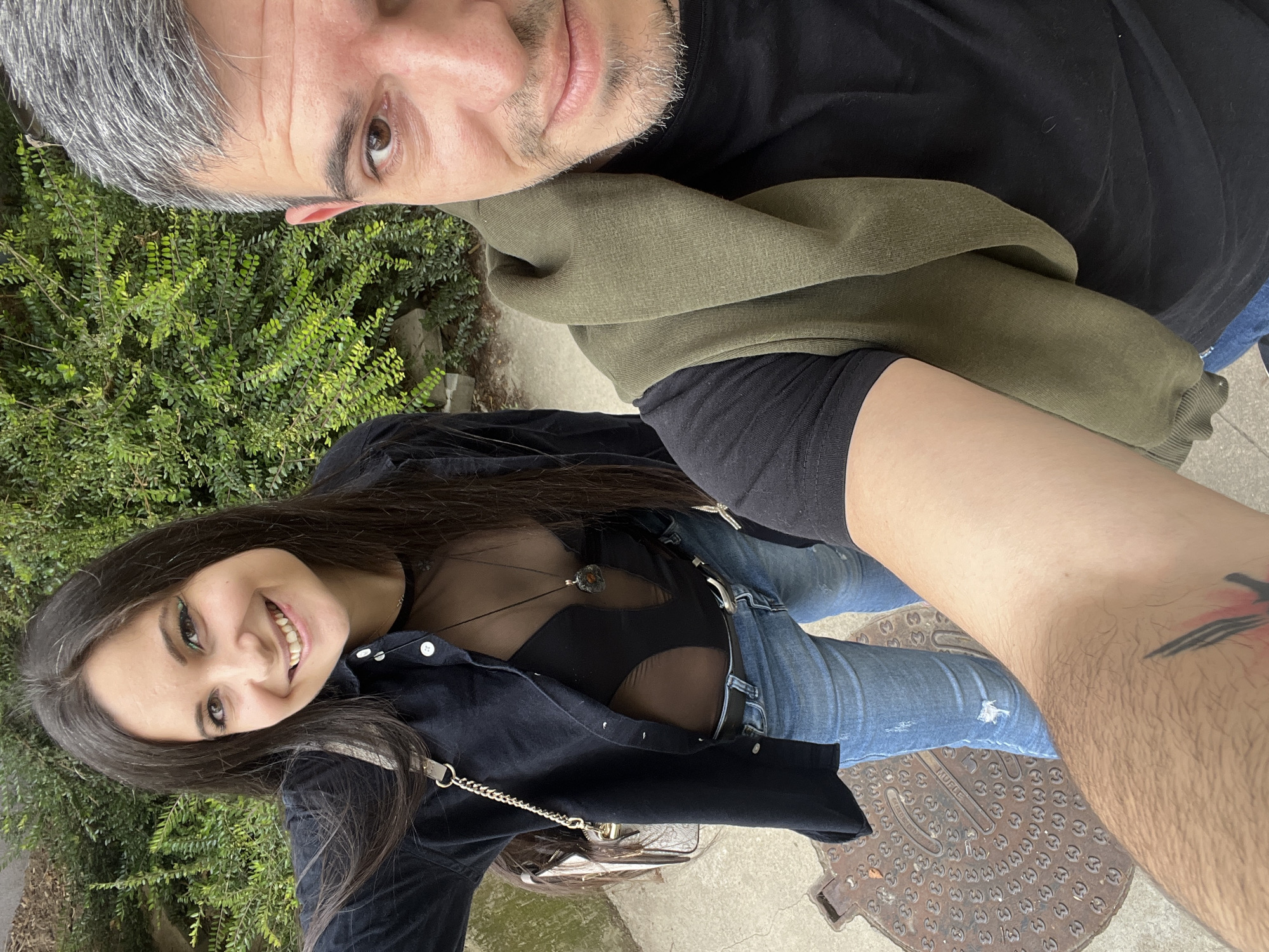 This is a selfie taken outdoors, likely on a sidewalk next to a dense green hedge. A woman with long dark hair and green eye makeup smiles at the camera, wearing a black open shirt over a sheer black top and blue jeans. A man with short graying hair and a beard, wearing a black t-shirt and a green hooded sweatshirt draped over his shoulders, is also visible in the frame, his arm extending towards the camera. A metal manhole cover is visible on the ground near the woman's feet. The scene suggests a casual outing in a city environment.