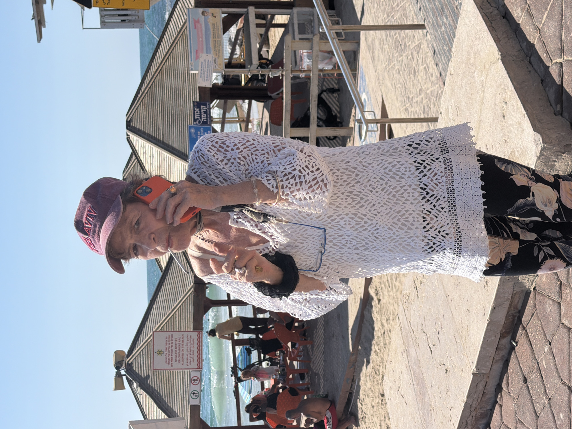 An elderly woman stands on a walkway, talking on a bright orange smartphone. She wears a burgundy baseball cap with "Tel Aviv" embroidered on the front, a white lace top, and floral print pants. She also wears glasses, a watch, bracelets, and rings. She is partially silhouetted.
Behind her is a beach scene, including a beach shelter, the ocean, and beachgoers. The beach shelter has signs in Hebrew and English that state that glass bottles or containers are not allowed on the beach.
The location is Tel Aviv, Israel.