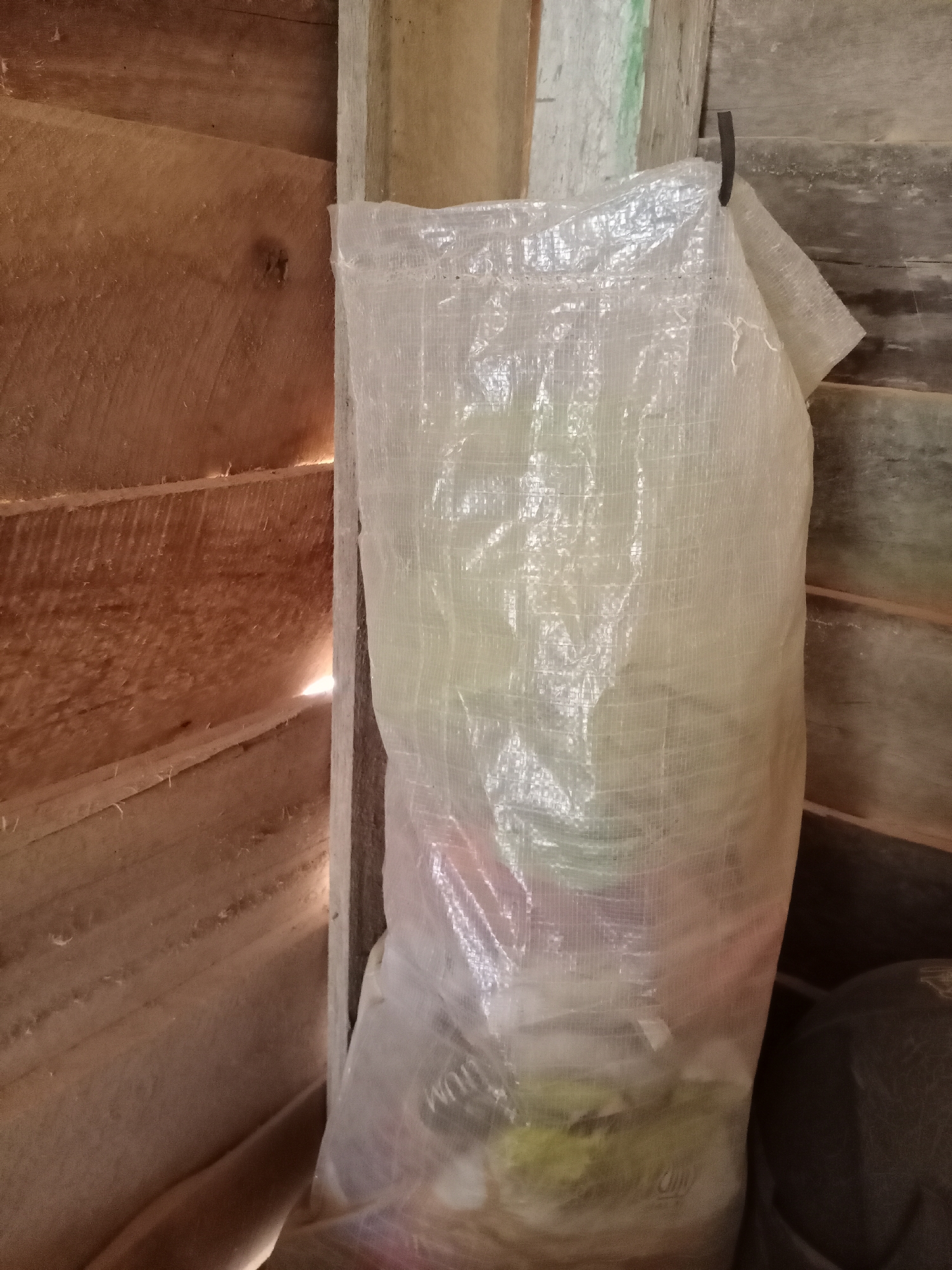 A clear plastic bag filled with what appears to be trash is leaning against a wooden structure. The bag is translucent, allowing some of the contents to be visible, including what looks like food waste and plastic. The wooden structure is made of rough-hewn planks, suggesting an outdoor or storage area. A dark object, possibly another bag or piece of furniture, is visible to the right of the plastic bag. The overall scene suggests a simple, possibly rural, setting in Ketapang, Indonesia, where such storage solutions might be common for waste disposal. The lighting is dim, with a shaft of light illuminating the back of the wooden structure, hinting at an opening or a gap in the construction.
