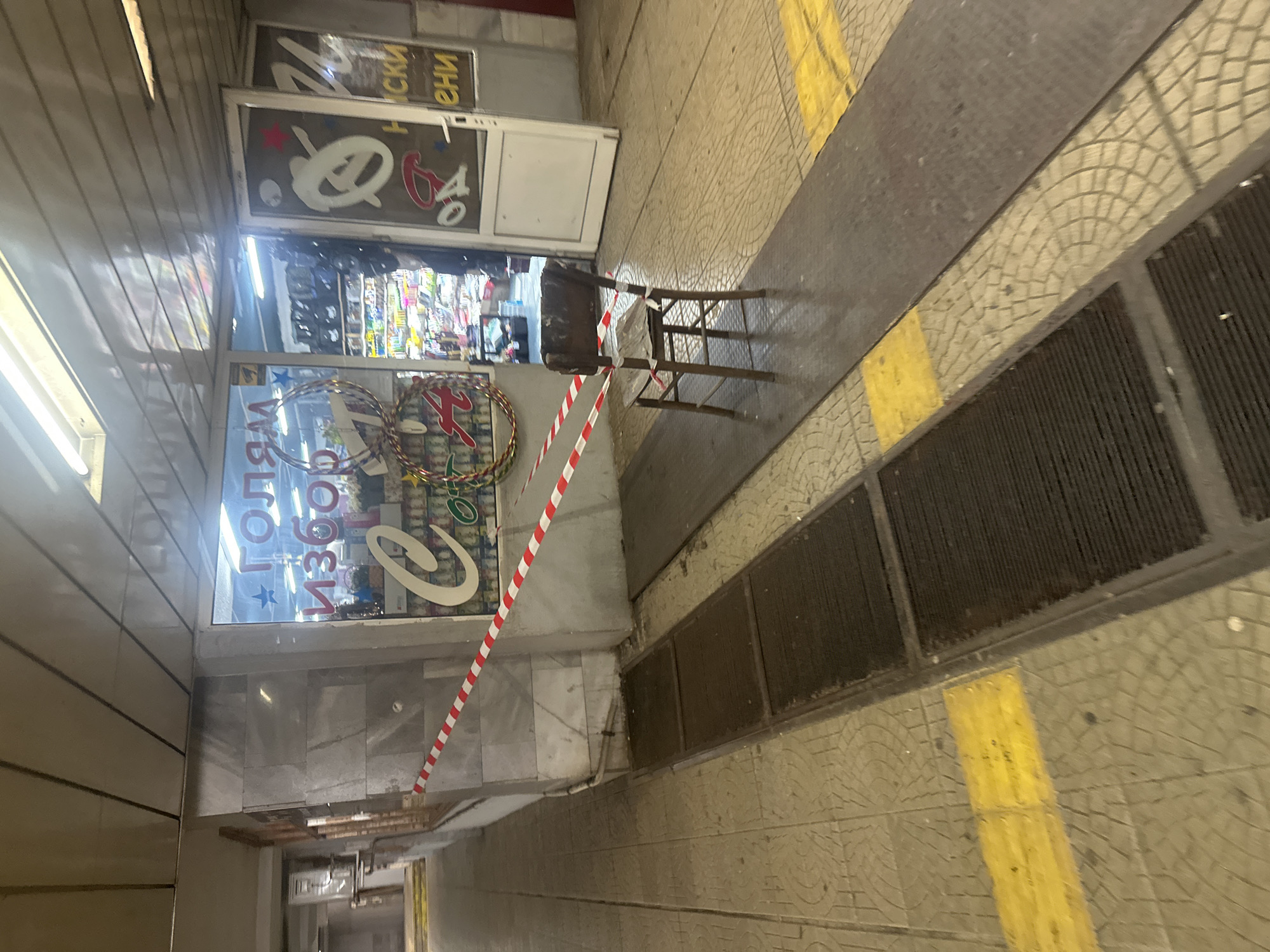 The image depicts a street-level view of a commercial establishment in Sofia, Bulgaria, likely a small shop or kiosk, cordoned off with red and white striped tape. The shop's interior is visible through a large glass window and an open doorway. The window displays some text, possibly in Cyrillic, including what appears to be "ГОЛЯМ ИЗБОР" (meaning "large selection"). Inside, shelves stocked with various items are visible, illuminated by fluorescent lighting. An old wooden chair is placed in front of the entrance, also partially obstructed by the tape. The floor is paved with patterned tiles, and there are metal grates or drain covers set into the pavement in the foreground. Yellow tactile paving is present on the floor, indicating accessibility features. The overall impression is one of a temporarily closed or inaccessible shop in an urban environment.