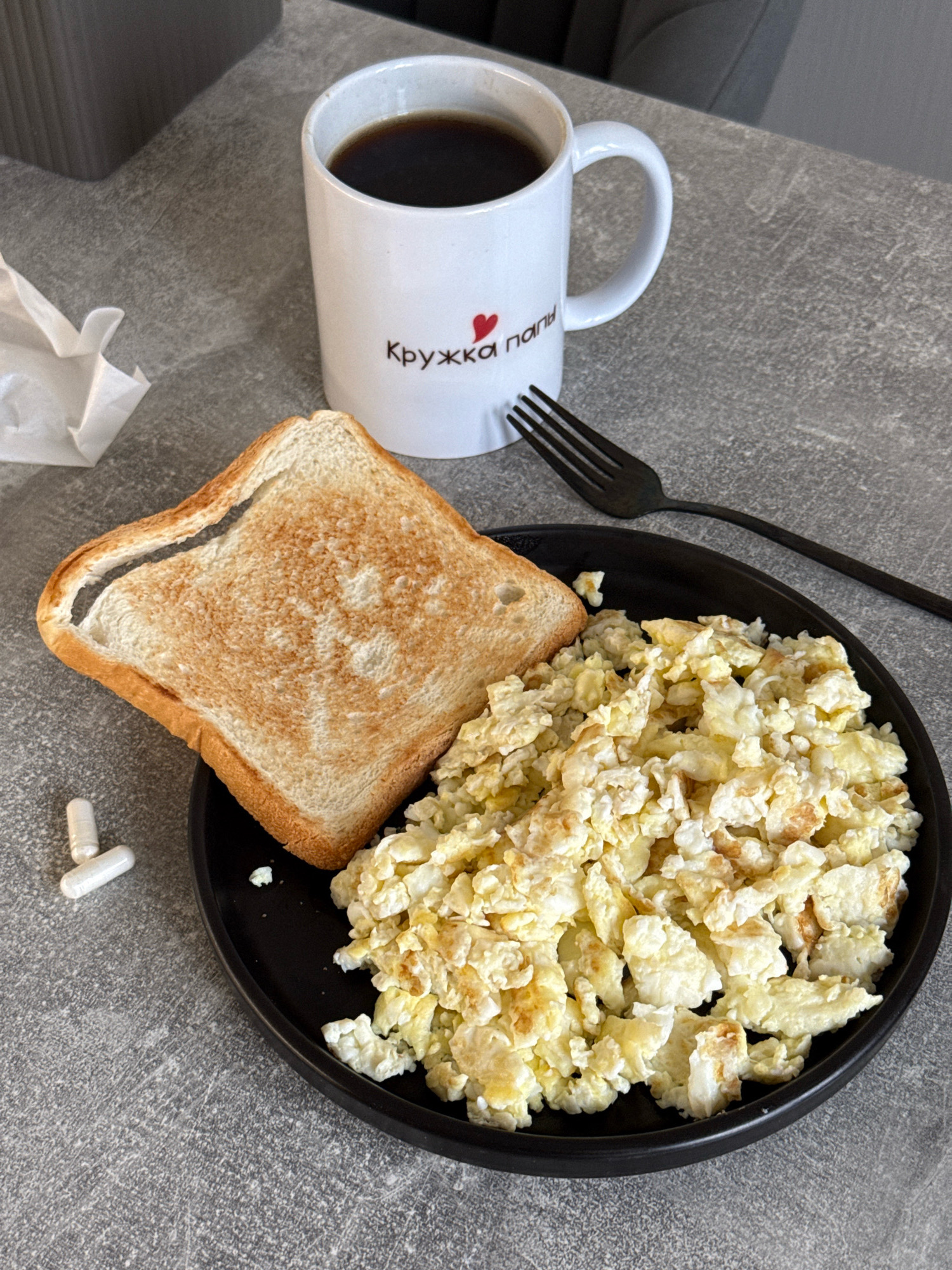 A breakfast scene is captured on a gray, speckled surface that resembles a countertop. In the upper portion of the image, a white mug filled with dark liquid, presumably coffee, sits slightly off-center. The mug has red lettering and a small red heart graphic. To the left of the mug, a crumpled white napkin is visible.
Dominating the center and lower part of the frame is a black plate containing a portion of scrambled eggs and a slice of toasted bread. The scrambled eggs are fluffy and appear lightly browned in places. The toast is golden brown with a slightly darker crust. To the left of the plate, two white capsules or pills are placed on the gray surface. A black fork rests on the plate, positioned above the scrambled eggs and toast. The overall impression is of a simple, home-style breakfast. The location is unknown.