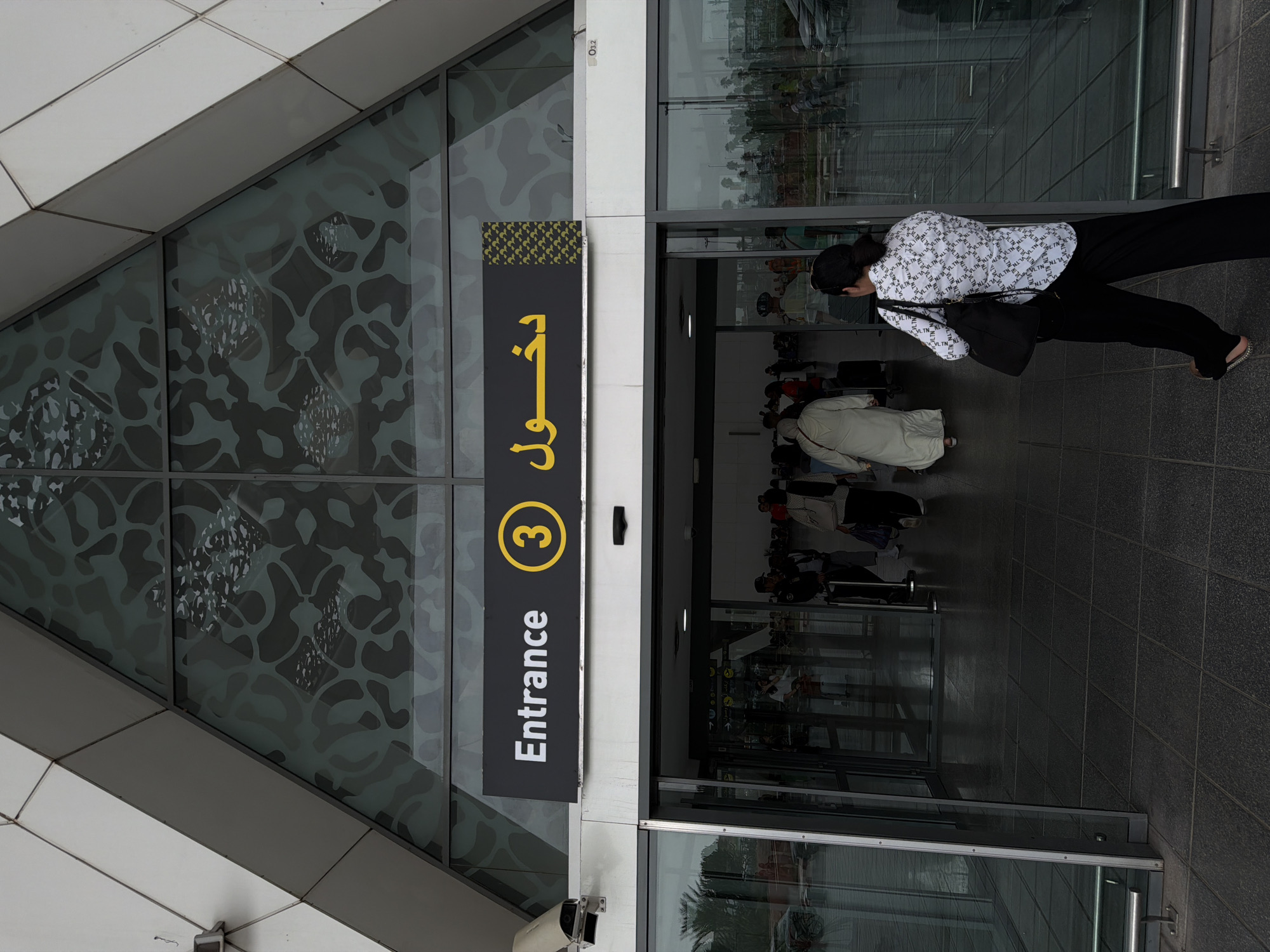 The image shows the entrance to a building, likely an airport or large public facility, with signage in English and Arabic. The English text reads "Entrance 3". The Arabic text translates to "Entrance". A woman wearing a white shirt with a repeating pattern and black pants is walking away from the camera and towards the entrance. She is carrying a black bag over her shoulder. In the background, several people are seen entering the building, some with luggage. The building's architecture features a large triangular glass facade with a patterned design. The scene appears to be outdoors in daylight. Based on the signage and general architectural style, this could be the Marrakesh Menara Airport in Marrakesh, Morocco.