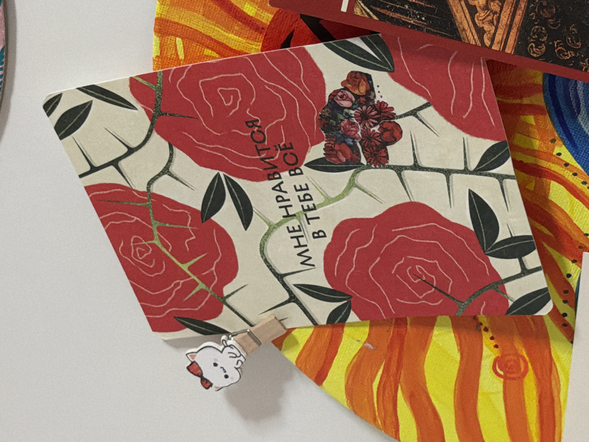 A close-up shot shows a postcard with a floral design featuring red roses and green thorny stems. The postcard is angled and partially overlaps other colorful artwork. Text on the postcard, written in Cyrillic, reads "МНЕ НРАВИТСЯ В ТЕБЕ ВСЁ," which translates to "I like everything about you." A small, wooden clothespin with a cartoon cat design is attached to the top left corner of the postcard. The background is a plain white surface, suggesting an indoor setting. The overall scene appears to be a display of art or personal mementos. There is no information in the image to suggest a specific location in Moscow, Russia.
