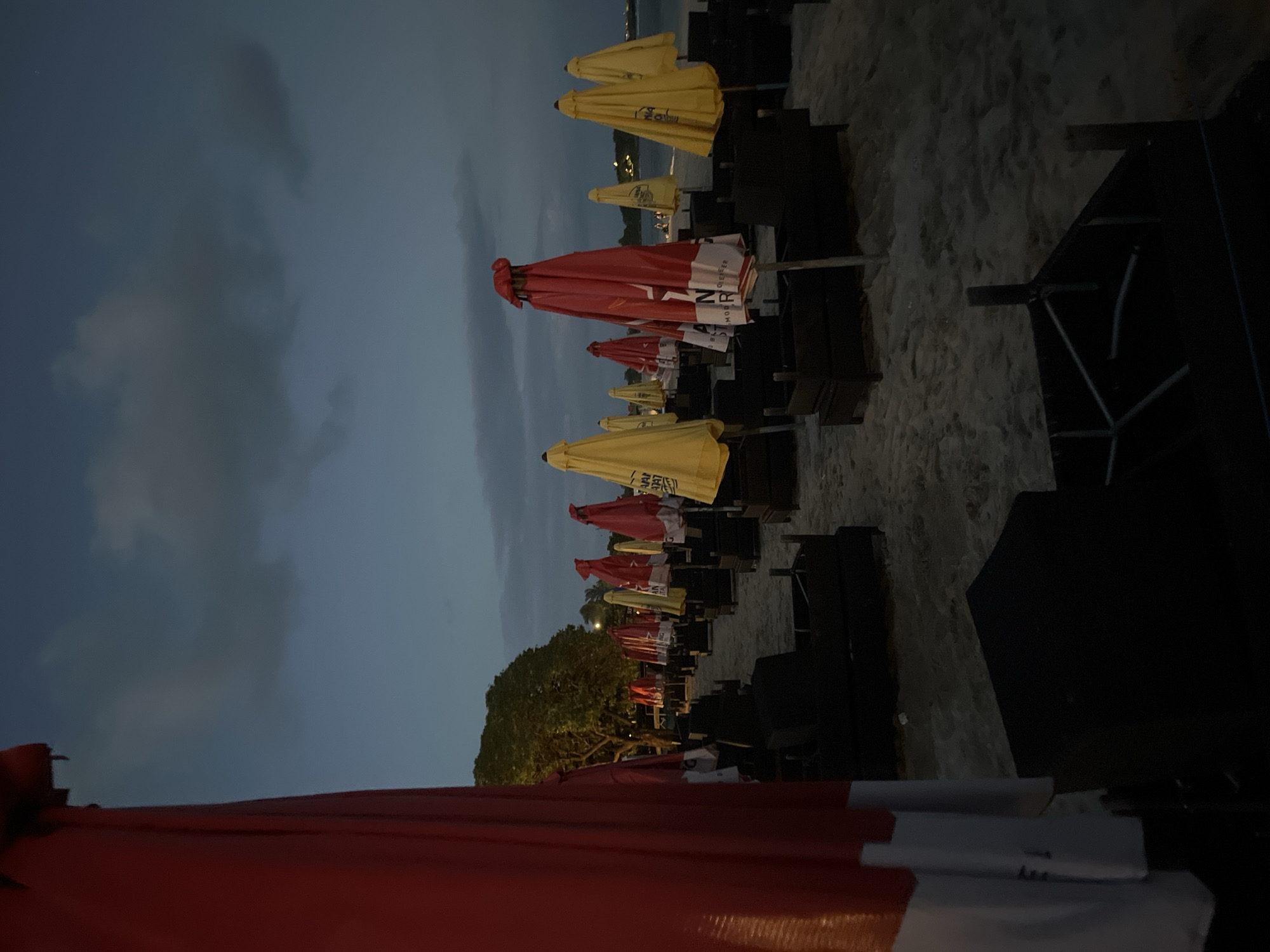 The image shows a line of closed beach umbrellas and lounge chairs on a sandy beach. The umbrellas alternate between red and yellow. In the background, there are trees and a body of water. The sky is overcast. The location is Kuta Selatan, Indonesia. The image appears to have been taken in the evening or at dusk. There are no people visible in the frame.