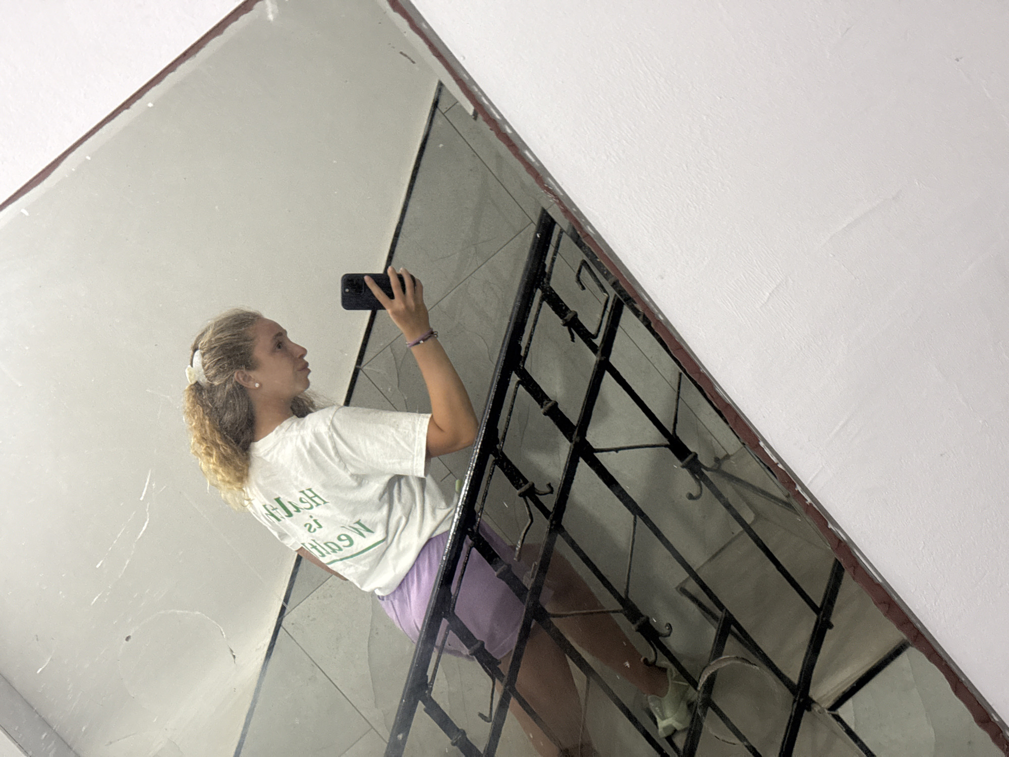 A young woman with blonde curly hair tied back in a scrunchie takes a mirror selfie in what appears to be a stairwell. She is wearing a white t-shirt with green lettering and light purple shorts. The background of the mirror reflection shows tiled walls and a black metal railing. She is holding a black smartphone in her right hand and appears to be looking at the phone's screen. Her left arm is resting on the railing. The woman is positioned on a staircase, with her body angled towards the mirror. The environment suggests an indoor setting, possibly an apartment building or public space. The image was taken in Tel-Aviv, Israel.