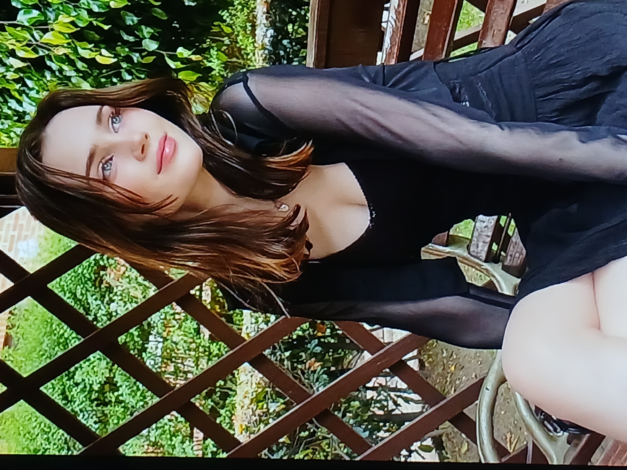 A young woman with long brown hair and blue eyes is sitting on a wooden bench. She is wearing a black dress with sheer sleeves. She is looking to her right with a slight smile. The background is a wooden lattice fence with green foliage behind it. There is no indication of this scene being located in Dnipro, Ukraine.