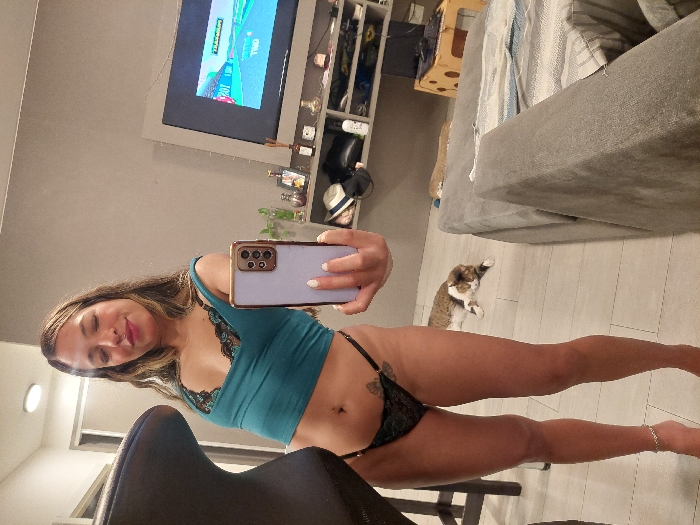 An adult female is depicted standing in a residential interior, consistent with a modern apartment in Buenos Aires, Argentina. She is facing forward, taking a selfie with a light-colored smartphone held in her right hand. The subject has light-toned skin, long wavy light-brown hair, and exhibits a subtle smile with eyes partially closed. She wears a teal-colored crop top with black lace trim and black lace thong underwear. A small tattoo is visible on her left upper thigh, and a silver-colored anklet adorns her left ankle. Her fingernails are painted a light, possibly white, color.
The background features a light grey wall and light-toned tile flooring. A dark, upholstered chair is partially visible on the far left. A grey, upholstered sofa with a striped throw blanket occupies the right side of the frame. In the center-rear, a flat-screen television is mounted on a grey entertainment unit, displaying a digital interface that includes the text "20Y" and "TRAGETORIO." Below the television, shelving units contain various decorative objects such as bottles, plants, and headwear. A domestic calico cat is lying on the floor in the mid-ground, positioned between the female subject and the sofa. A light-colored wooden structure, possibly a cat bed or furniture, is partially visible behind the sofa.
