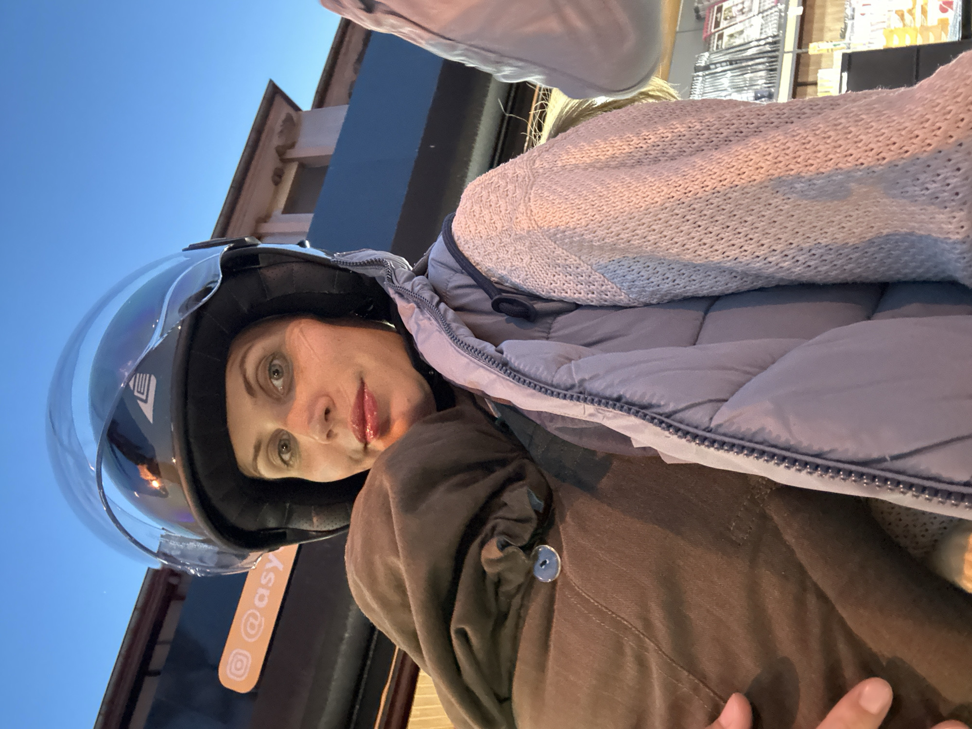 A woman wearing a motorcycle helmet with a clear visor is facing the camera. She is also wearing a grey padded jacket, a light grey knitted cardigan and a brown jacket. A sticker with "@asy" and an Instagram logo is attached to the helmet. The background includes a building with architectural details, a store with items on shelves and the evening sky. The location is likely a commercial area in Fethiye, Turkey.