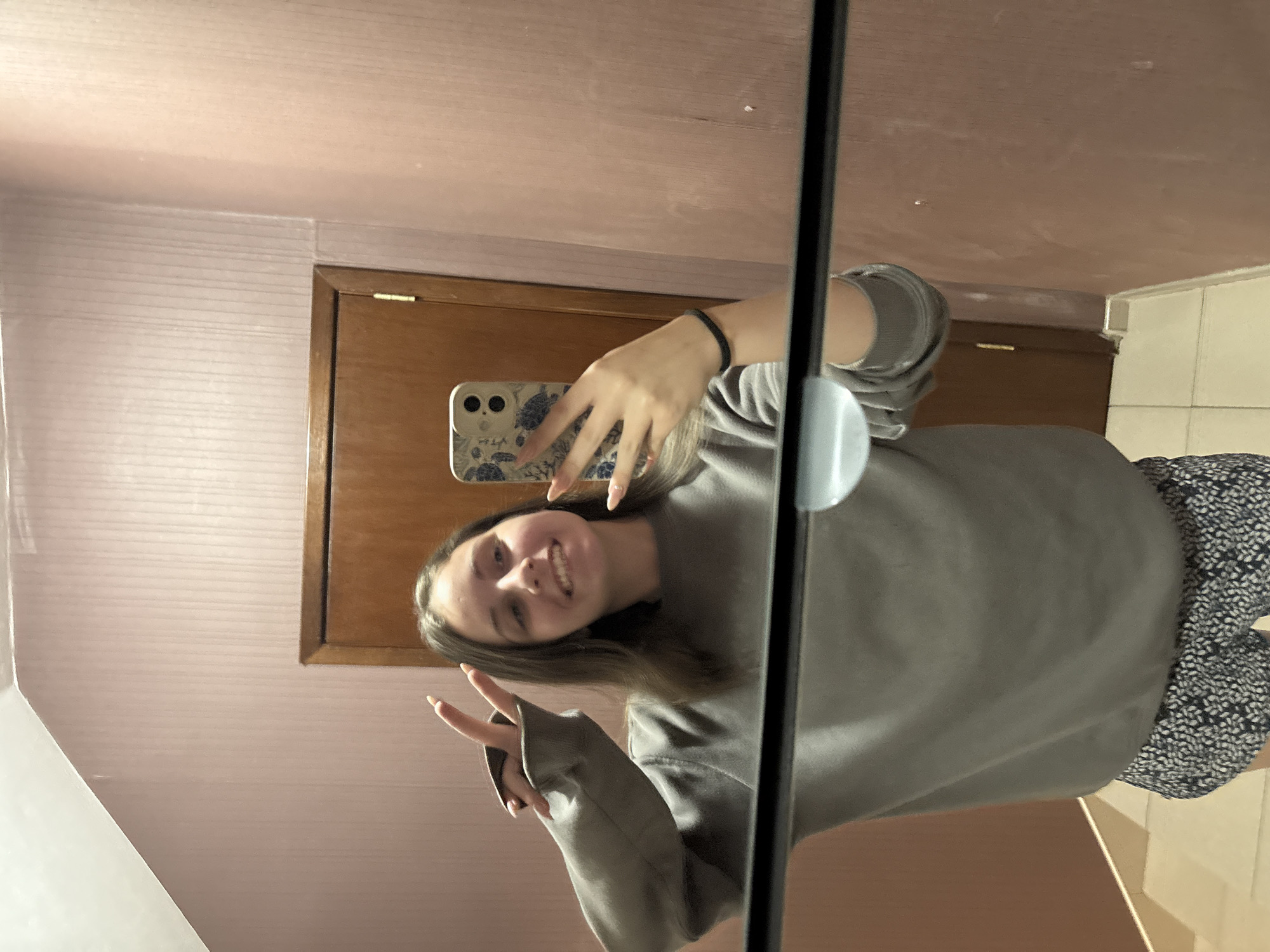 A young woman with long, light brown hair, wearing a grey sweatshirt and patterned shorts, stands in front of a mirror, taking a selfie with her phone. She is smiling and holding up two fingers in a peace sign. The background shows a pink, vertically striped wallpaper and a wooden door. The scene appears to be indoors, possibly in a residential setting. The image was taken in the city of Bulgarovo, Bulgaria.