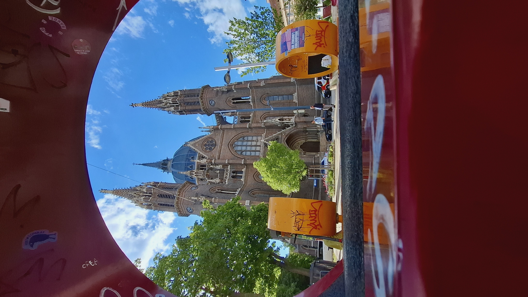 The image shows a church with a blue dome and two tall spires, framed by what appears to be a public art installation or street furniture. The church is located in Vienna, Austria. The sky is blue with scattered clouds. In the foreground, there are orange cylindrical structures with graffiti. People are seen walking on the sidewalk next to the church. There is a red object with stickers in the far foreground. The orientation of the image is rotated.
