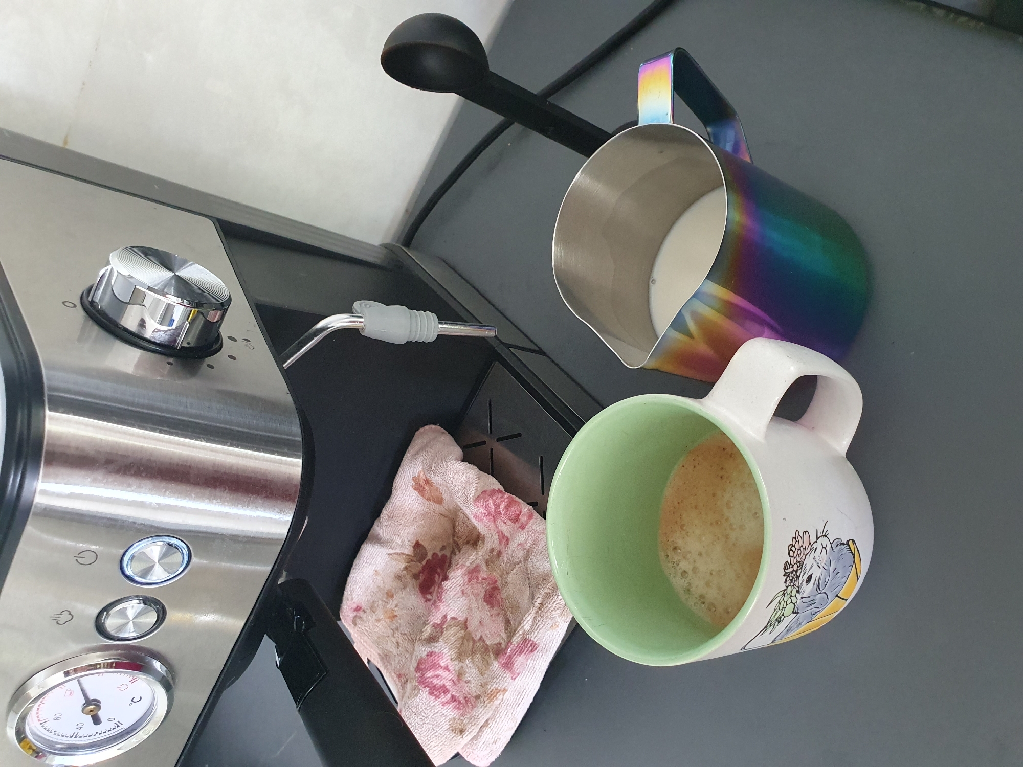 An espresso machine is shown in the background of a dark gray countertop. To the left of the machine is a small, white towel with a floral pattern. In the foreground, a white mug with a drawing of a rabbit is filled with a foamy, light brown liquid. Next to the mug is a rainbow-colored stainless steel milk pitcher filled with white liquid. A black scoop is resting on the countertop behind the milk pitcher.