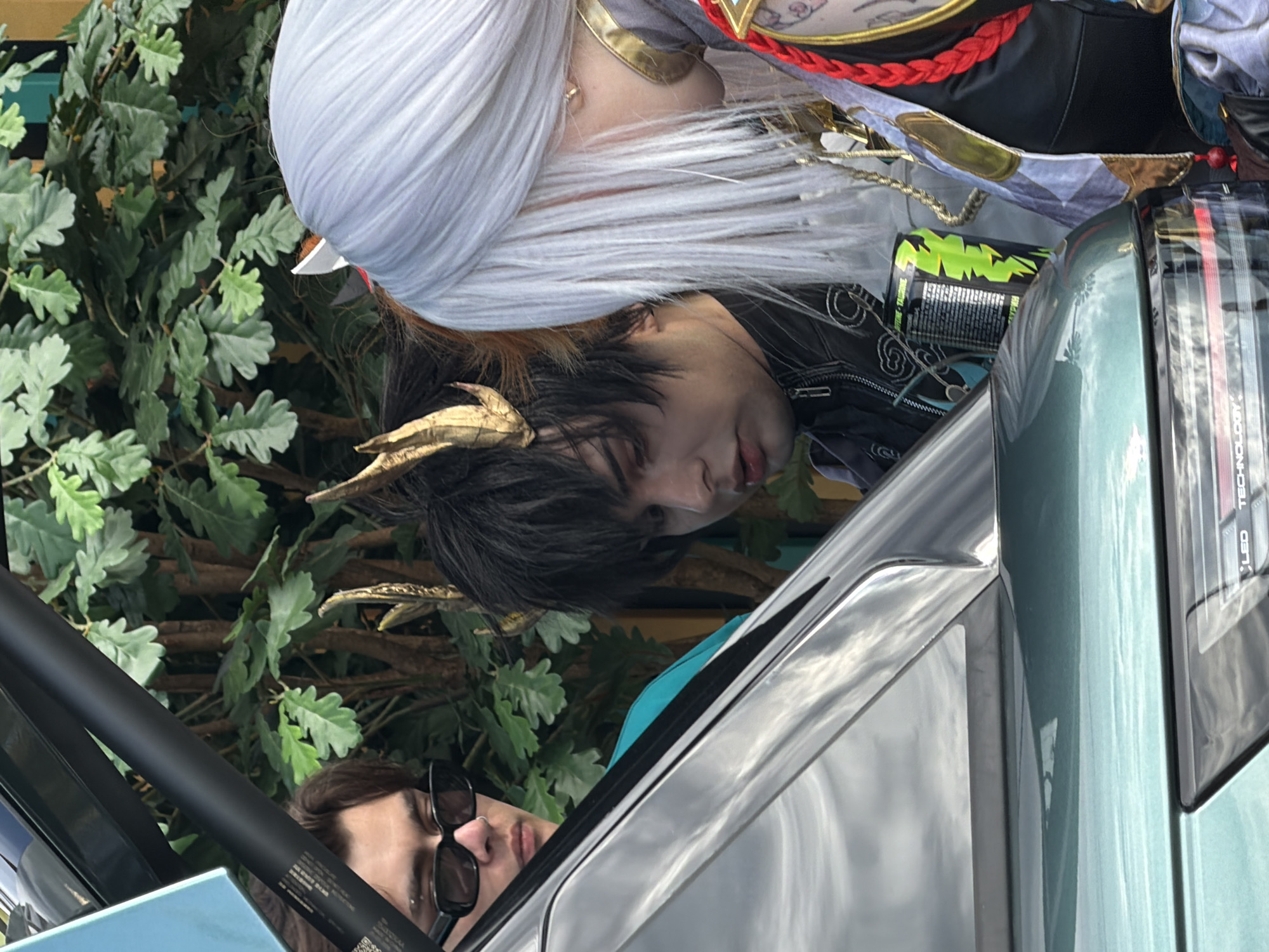 Three individuals, two appearing to be cosplayers with elaborate costumes and wigs, are gathered near the open trunk of a dark-colored vehicle, possibly a car. The person on the left is wearing black sunglasses and a light blue shirt. The person in the middle has dark hair with gold, horn-like accessories and is looking down. The person on the right has long, white hair and is also looking down, wearing a detailed costume with gold and red accents. A green energy drink can is visible between the middle and right individuals. The background consists of lush green foliage, possibly from trees or bushes. The scene appears to be outdoors, with natural light. The car's trunk is open, revealing a portion of its interior and the surrounding environment. The vehicle's paint has a reflective surface, showing a cloudy sky. The overall setting is casual and appears to be a gathering or event. The location is Saint Petersburg, Russia.