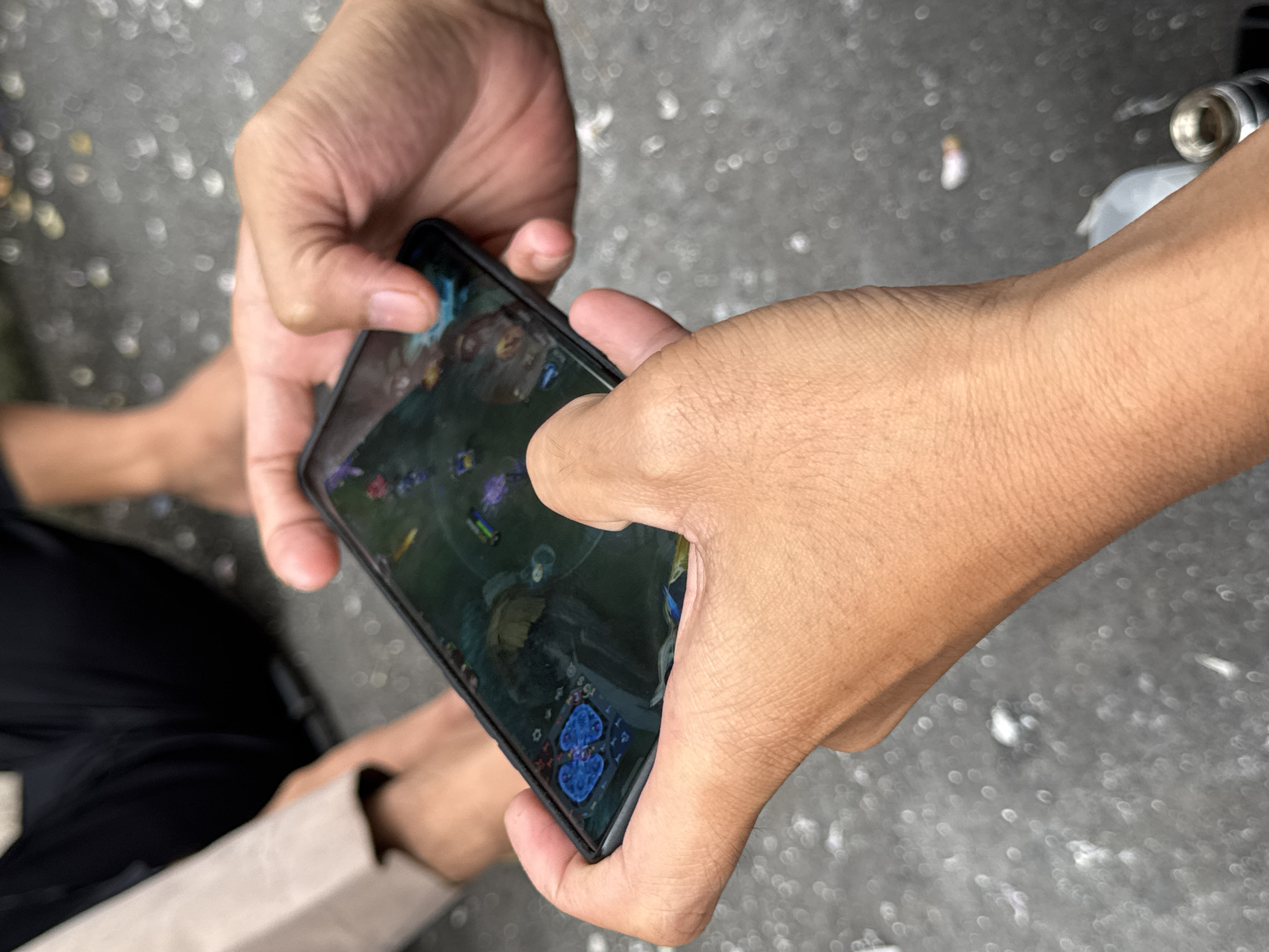The image shows a close-up of two pairs of hands holding and interacting with a smartphone displaying a video game. The hands are tanned and appear to be those of young adults. The phone screen shows a fantasy-themed game with colorful characters and a map interface. In the background, partially visible, are the legs and feet of two individuals, one wearing dark pants and the other bare-legged. The environment appears to be outdoors, possibly a street or public area, with a grey, textured ground covered in small white and light-colored debris. The overall impression is one of casual interaction and entertainment. Given the style and common usage of mobile gaming, this scene could be taking place in a variety of urban or semi-urban settings, and without further information, it is impossible to pinpoint the exact location within Kuta Selatan, Indonesia.