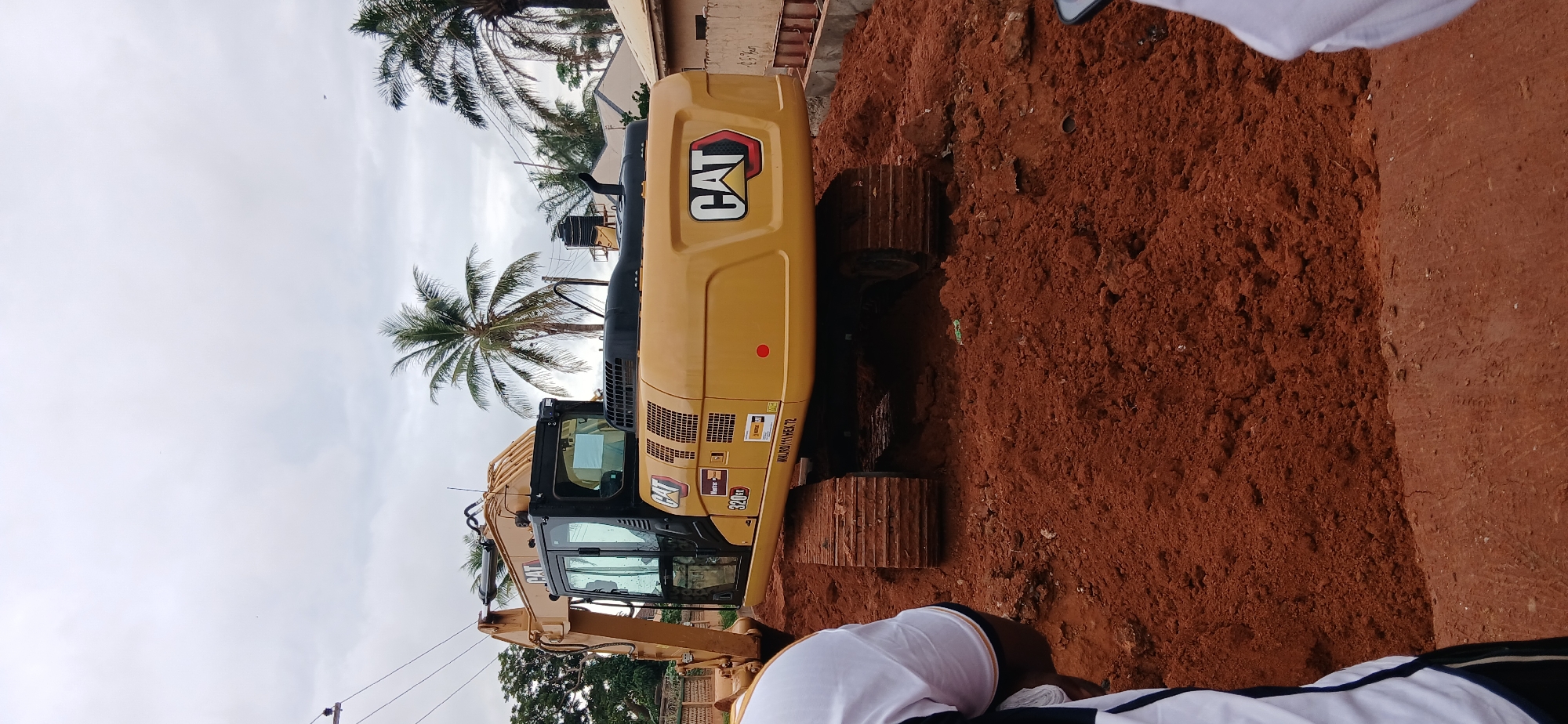 In Sabon Tasha, Nigeria, a yellow Caterpillar 320EX excavator is positioned on a mound of reddish-brown soil. The excavator, featuring a black cab with tinted windows, has the "CAT" logo prominently displayed on its rear. To the left of the excavator, part of a person wearing a white shirt with blue and orange trim is visible. In the background, palm trees and houses suggest a residential or semi-rural environment under an overcast sky. The ground is uneven, covered in soil, with some tracks from the excavator's treads visible. Electrical wires are strung overhead.