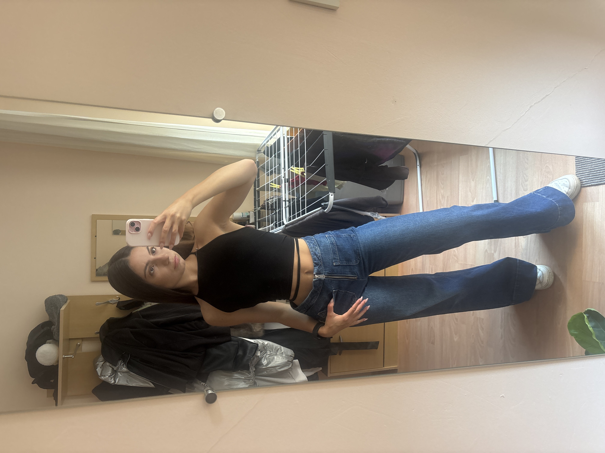 In a well-lit room, a young woman is taking a full-body mirror selfie. She is wearing a black crop top and high-waisted blue jeans. The jeans are wide-legged and feature a zipper closure and some detailing around the pockets. She has long, dark hair and is holding a pink smartphone in her right hand. Her left hand is resting on her hip, with her fingers splayed. She is looking at her reflection in the mirror, which occupies most of the frame.
Behind her, the reflection shows a closet with several items of clothing hanging on hangers and shelves. A white drying rack with some yellow clothes pegs is also visible. The room has light-colored walls and a wooden floor. A framed mirror is hanging on the wall, reflecting a portion of the room. A large green plant is partially visible in the lower right corner of the reflection. The overall impression is that of a casual, domestic setting, possibly a bedroom or dressing area. The lighting suggests it is daytime. There is no visible text in the image.