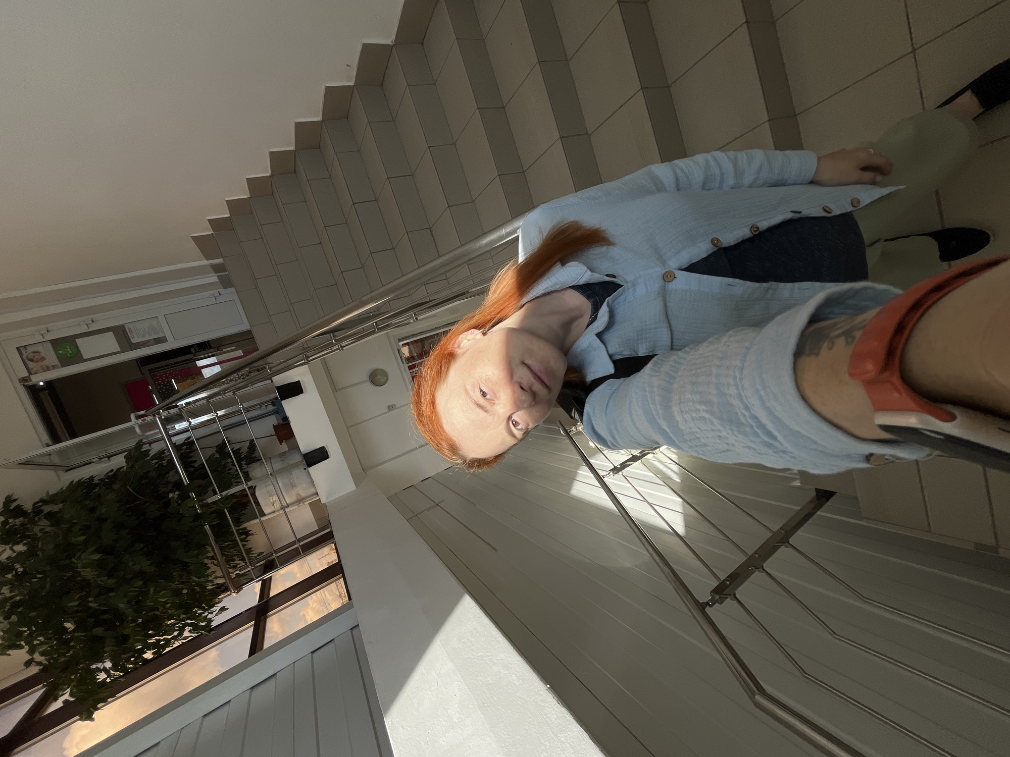 A woman with red hair is taking a selfie on a staircase. She is wearing a light blue button-up shirt over a dark shirt and light green pants. She has a backpack on her left shoulder and a smartwatch on her left wrist. The staircase has tiled steps and a metal railing. A large potted plant is visible near a window on the left side of the image, through which a sunset sky is visible. The scene appears to be indoors, possibly in a public building or an office. The image was taken in the city of Chaikovsky, Russia.