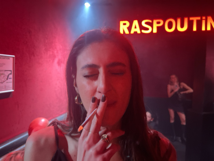 A woman with dark hair and closed eyes holds a lit cigarette near her lips, illuminated by a bright, overhead light. She wears a black top and multiple rings. The background features a red wall with an evacuation plan poster and the neon sign "RASPOUTIN." Another woman in a black dress and boots stands further back, partially obscured. The scene is awash in red lighting, suggesting a nightclub or bar atmosphere. A man is slightly visible behind the second woman.
