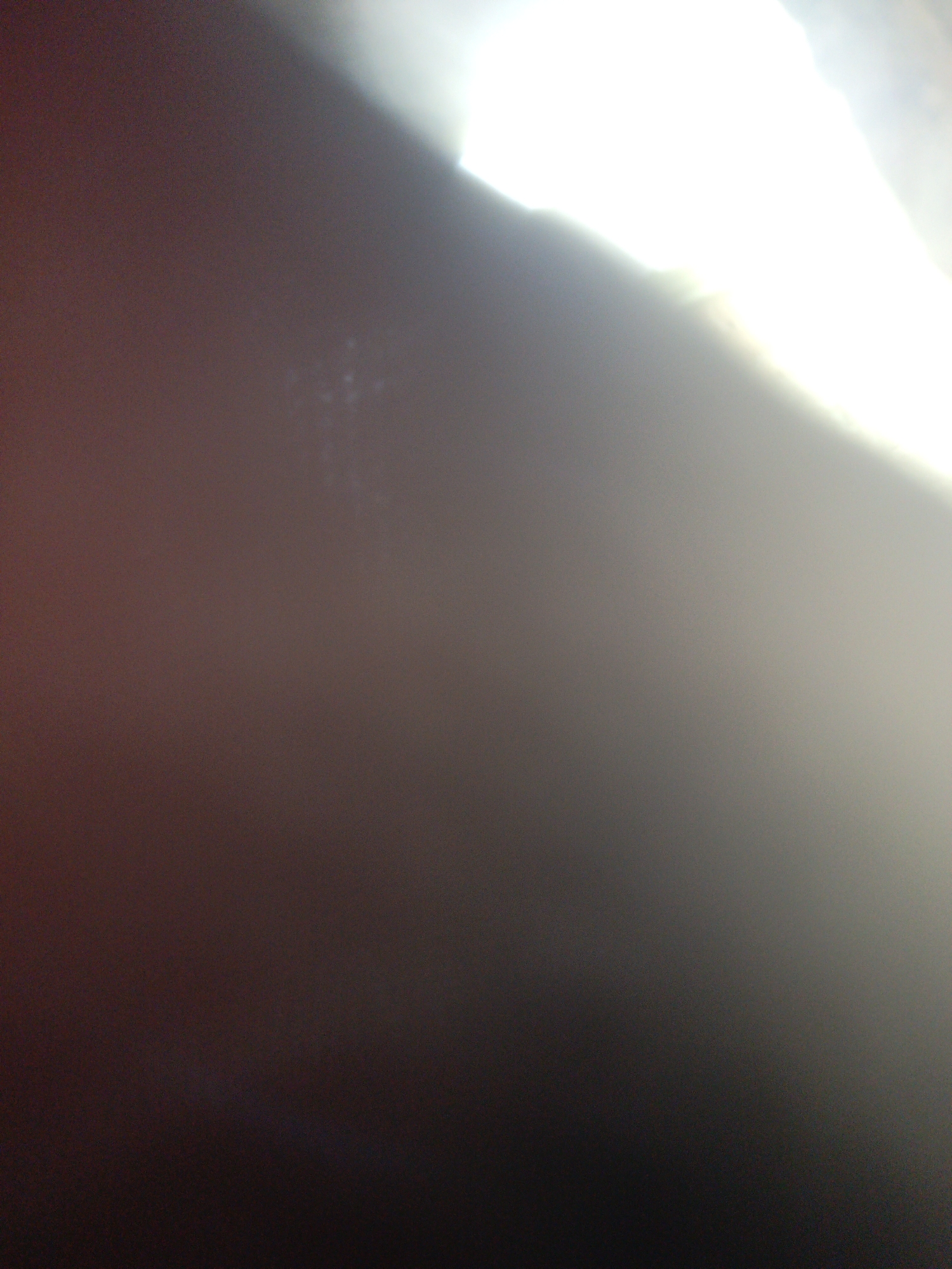 The image is extremely blurry and underexposed, making it difficult to discern specific details. The right upper corner of the image has a bright white area, which may be a light source. The rest of the image is a gradient of dark brown/gray to black, with very little detail visible. There is no discernible information about people, subjects, objects, actions, interactions, or the environment that can be extracted from the image. The location is unknown due to the lack of identifiable features.