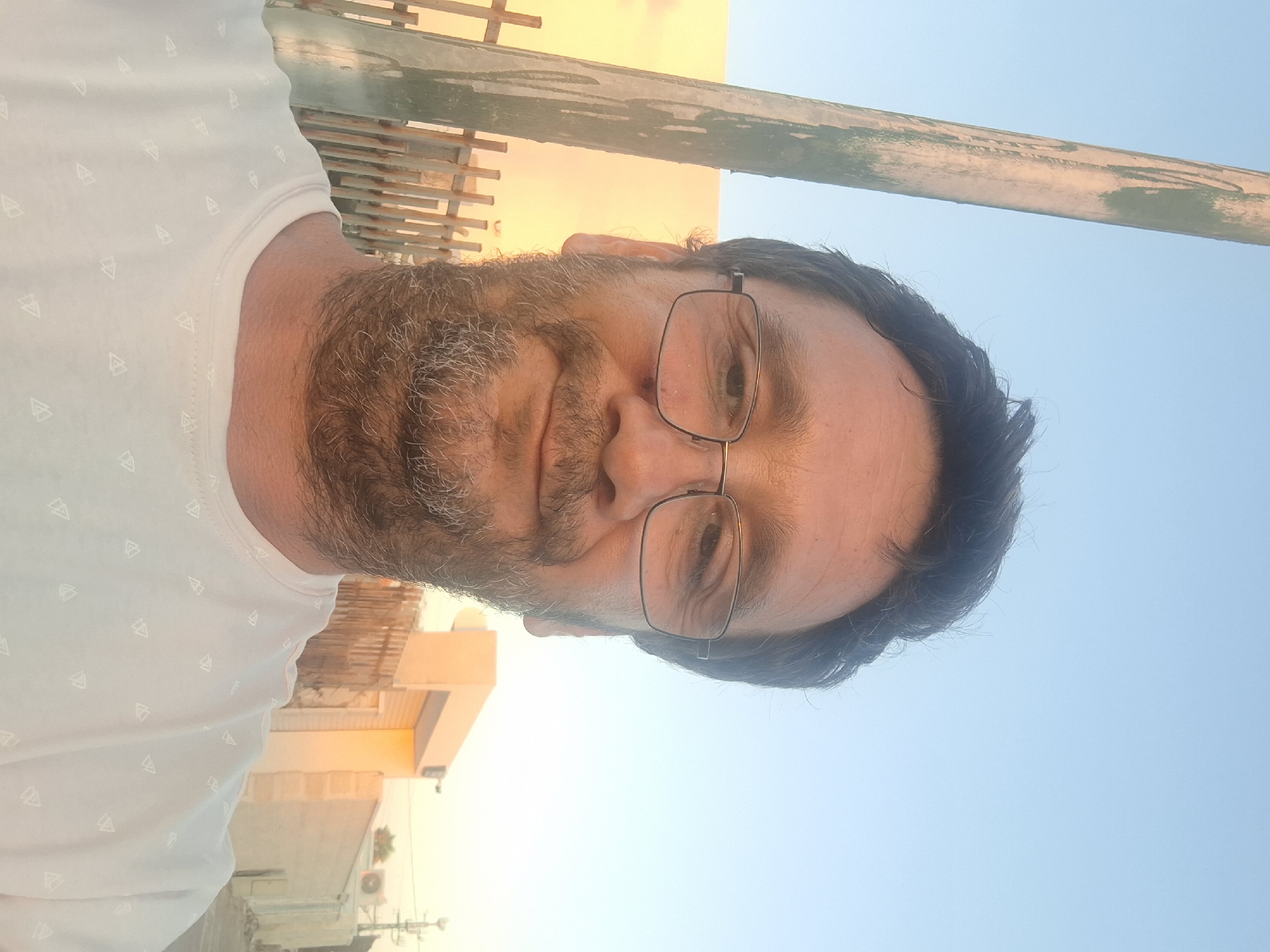 The image shows a man wearing glasses and a white t-shirt with a pattern of small triangles. He has dark, messy hair and a beard. He is looking at the camera and smiling slightly. The background is a blurred outdoor scene with a blue sky, a wooden pole on the left, and buildings. The lighting suggests it is either early morning or late afternoon, with warm, soft light illuminating the scene. The man appears to be in an urban or suburban environment.