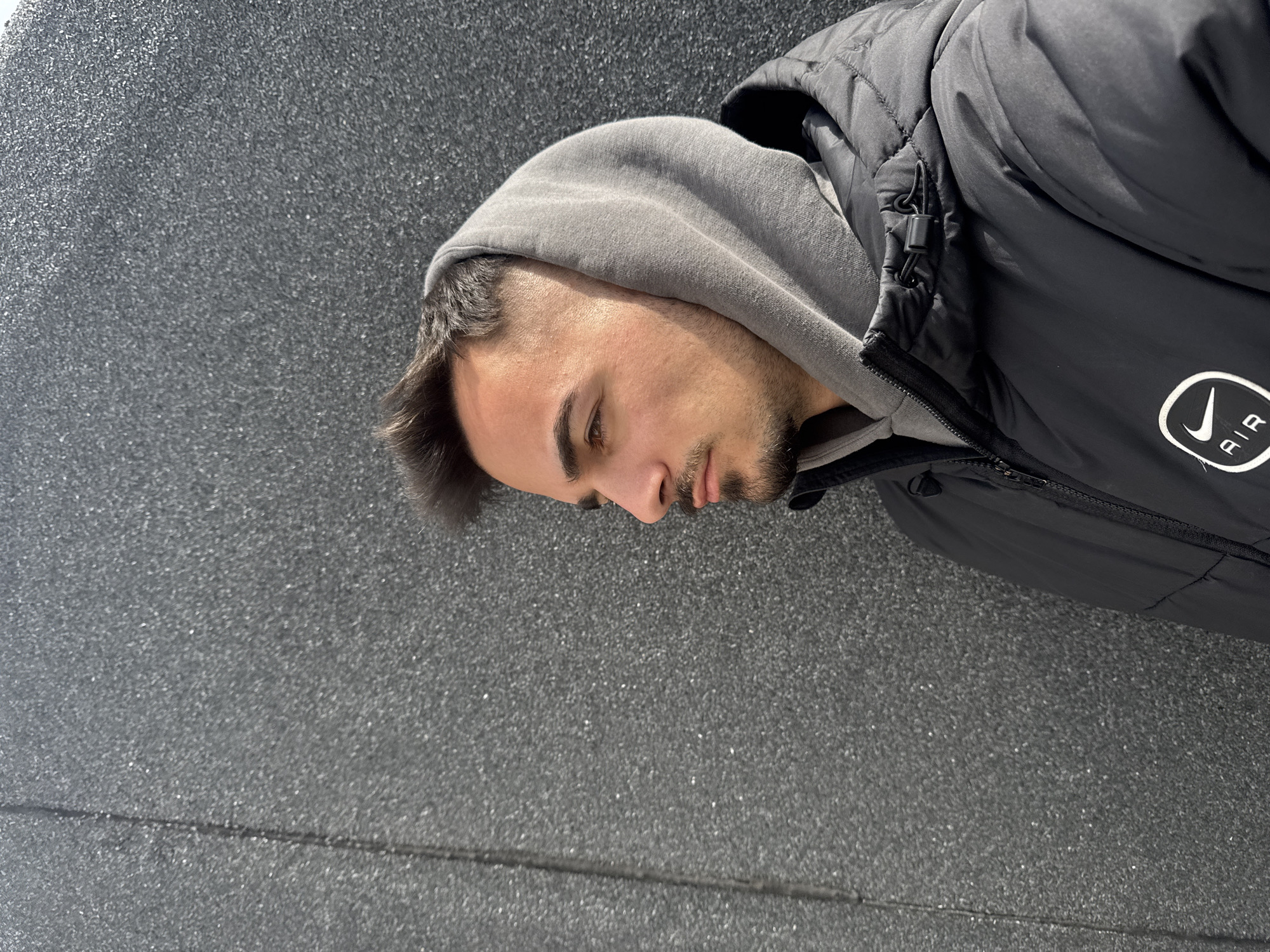 A young man stands with his head bowed, partially concealed within the hood of a grey sweatshirt. Over the sweatshirt, he wears a black padded jacket adorned with the Nike "Air" logo. The background is a grey textured wall. The location is undetermined; there is no identifying information within the image to establish the city of Sofia, Bulgaria.