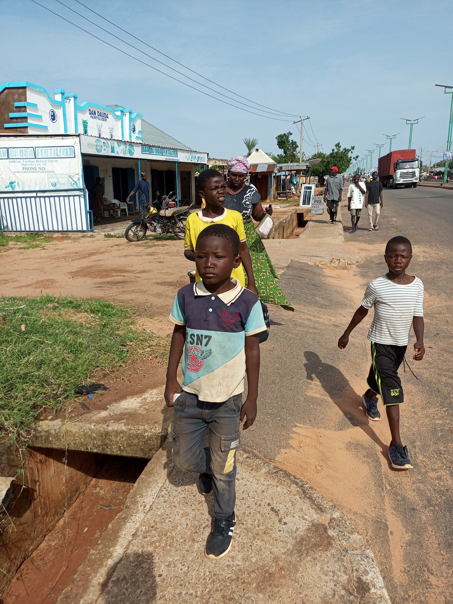 The image shows a street scene in Garko, Nigeria. In the foreground, two young boys are walking on a concrete path next to a ditch filled with reddish dirt and sparse grass. The boy on the left, wearing a multi-colored polo shirt and grey cargo pants, is walking towards the camera. The boy on the right, in a striped t-shirt and black shorts, is walking away from the camera. Behind them, a group of people are also walking on the sidewalk, including women in traditional attire and young men. A motorcycle is parked near a building on the left, which appears to be a shop or business with the sign "DAN DAUDA FERTILIZER". The street is paved, and there are streetlights and power lines visible. A red truck is driving in the distance on the right side of the road. The sky is clear and blue.