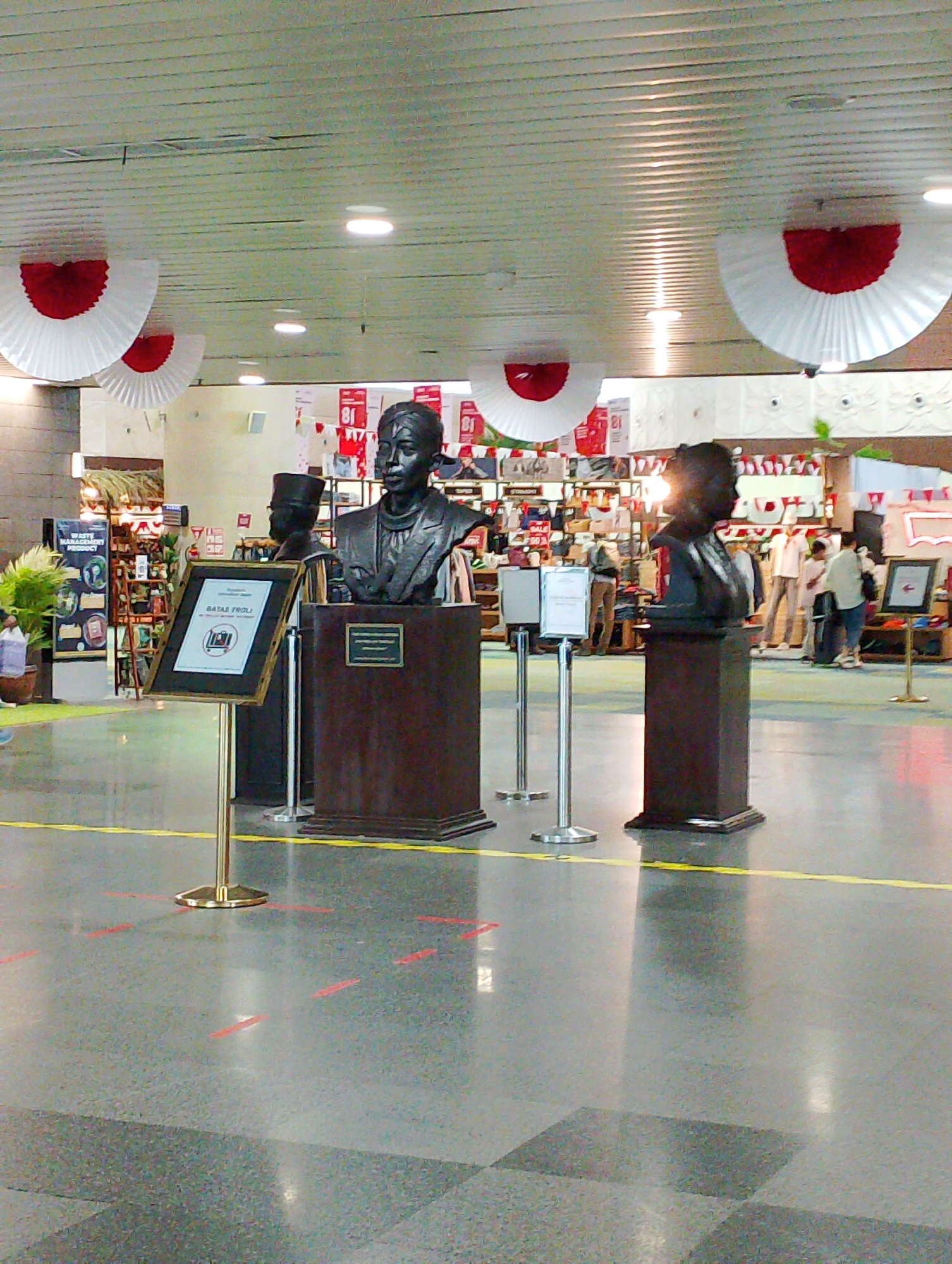 The image shows a public indoor space, likely a mall or a cultural center, decorated with red and white festive elements, possibly for a national holiday given the colors. In the foreground, there are two busts on pedestals. The bust on the left is partially obscured by a framed sign. The bust on the right is more prominent and is the primary subject. Behind the busts, there are display shelves filled with merchandise, and several people are visible browsing or walking through the area. The floor is a polished gray surface with yellow and red markings, suggesting a designated walkway or social distancing measures. The lighting appears to be from overhead fluorescent fixtures. The overall scene depicts a commercial or recreational environment.