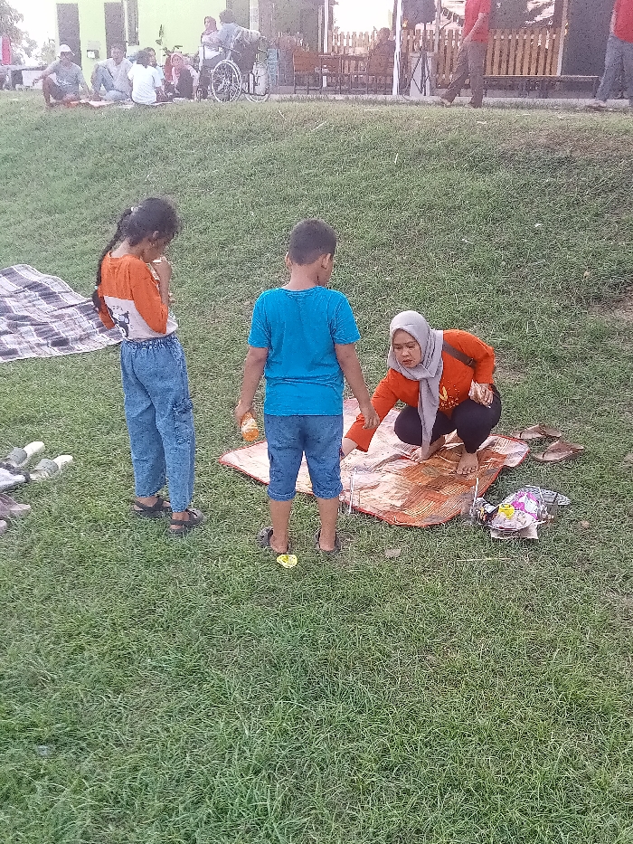 The image depicts a grassy area in front of a building with people sitting on a blanket. The coordinates -6.758, 111.075 place the location in the city of Jakenan, Indonesia. There are three people on the blanket; one adult woman is squatting on the ground, her arms outstretched towards the two children standing in front of her. The woman wears an orange and white top with a grey hijab, and the boy wears a blue shirt and blue shorts, while the girl wears an orange shirt and blue jeans. There is trash near the blanket. There are other people in the background, and a bicycle is visible in the upper left corner. The image is taken from a slightly elevated perspective and the light is bright.