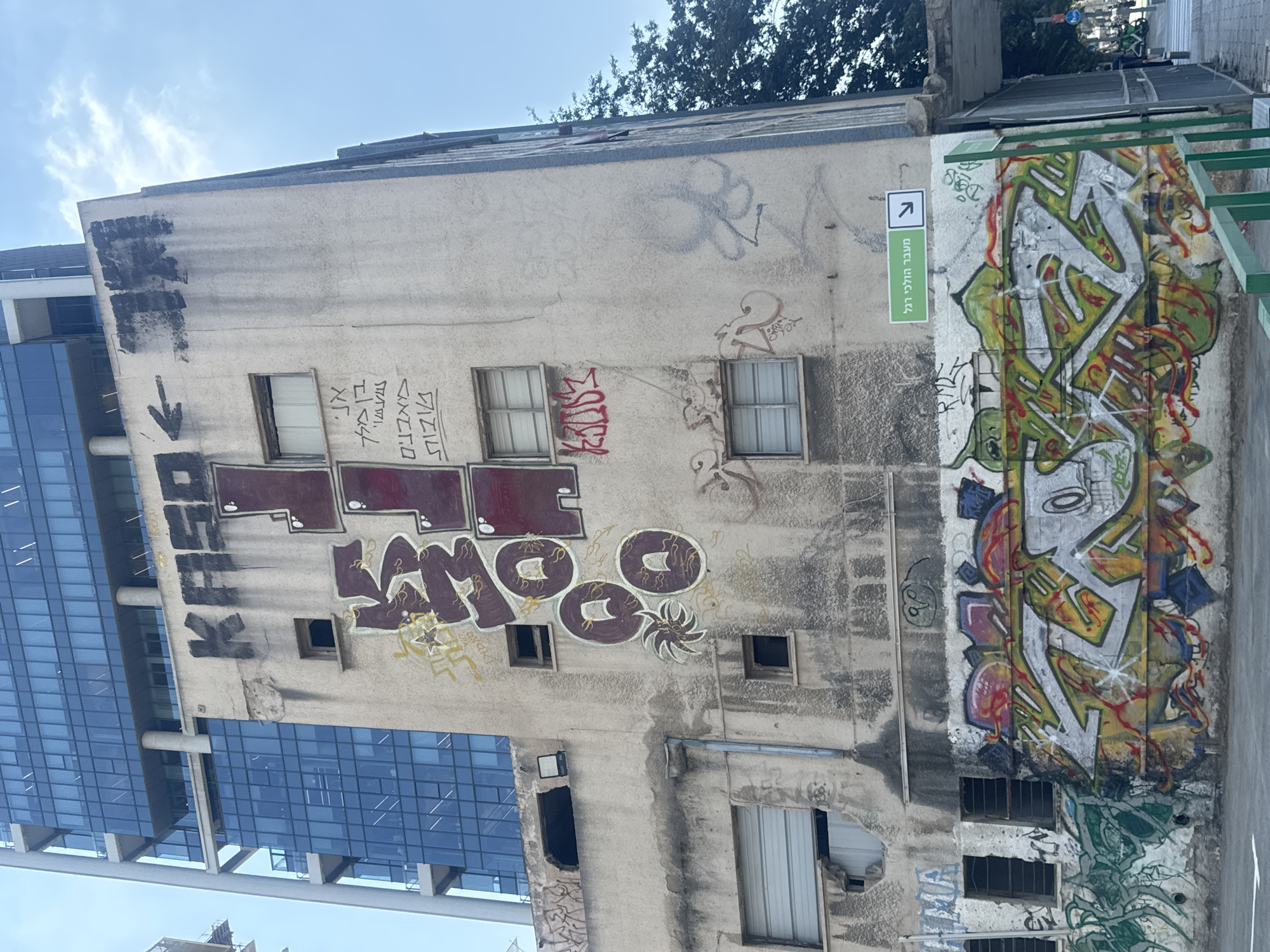 The image shows a weathered, multi-story building in Tel Aviv, Israel, covered in graffiti. The building is a mix of old concrete and a modern glass skyscraper is visible to the left. The graffiti ranges from large, bold lettering in dark red and black, to more intricate designs and tags in various colors. Some of the graffiti appears to be in Hebrew. One prominent piece of graffiti in dark red letters spells out "KASO" at the top of the building. Below it, there is more graffiti in Hebrew and in stylized lettering. At the base of the building, a large, colorful mural features cartoonish characters and abstract designs. A green sign with Hebrew text and an arrow points to the right, indicating a pedestrian crossing. The sky is clear and blue. There are no people visible in the immediate foreground, but a street with some parked bicycles is visible in the background on the right.