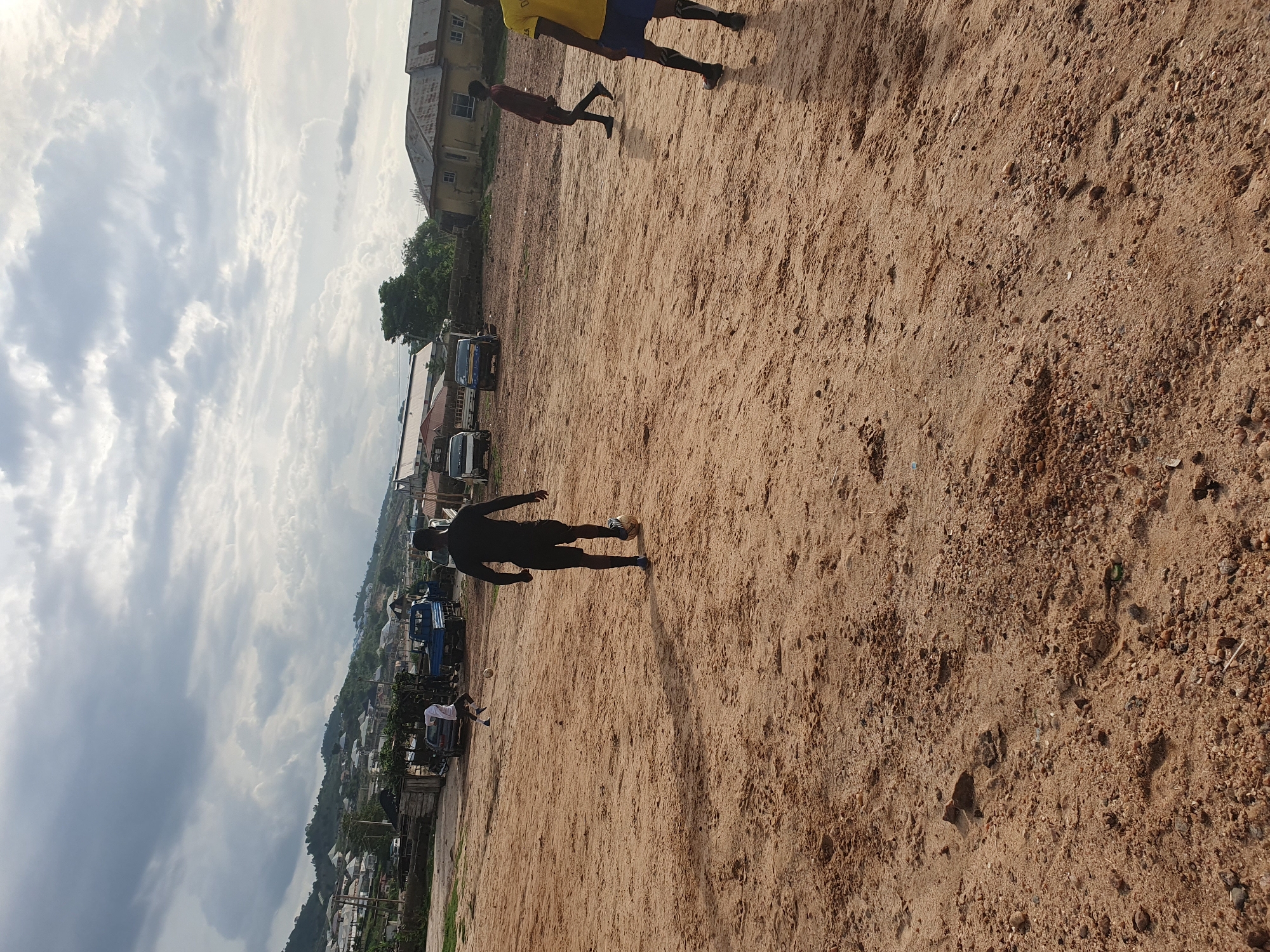 The image shows a dirt field in Lokoja, Nigeria, with at least three individuals visible. One person, dressed in all black, is walking across the field. Two others are partially visible at the top right, one wearing yellow. In the background, there are buildings, trees, and several vehicles parked along the edge of the field, and a view of a hillside with housing. The sky is overcast with clouds.