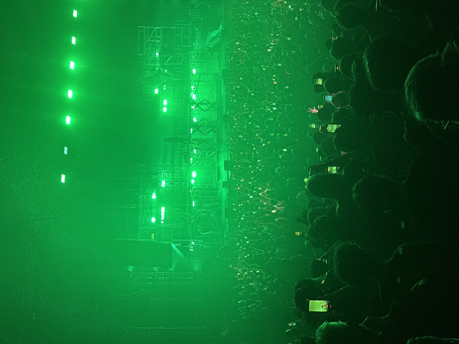 An indoor concert venue in Paris, France, is illuminated by pervasive green lighting. A vast audience occupies the foreground and midground, largely silhouetted, with many individuals holding smartphones vertically, their screens glowing green as they record the performance. On the stage in the background, a multi-level scaffolding structure is visible, adorned with numerous rectangular green light fixtures. A single figure, likely a performer, stands on an elevated platform at the center of the stage. Other indistinct figures are present on lower stage levels. The atmosphere contains visible haze, diffusing the green light.