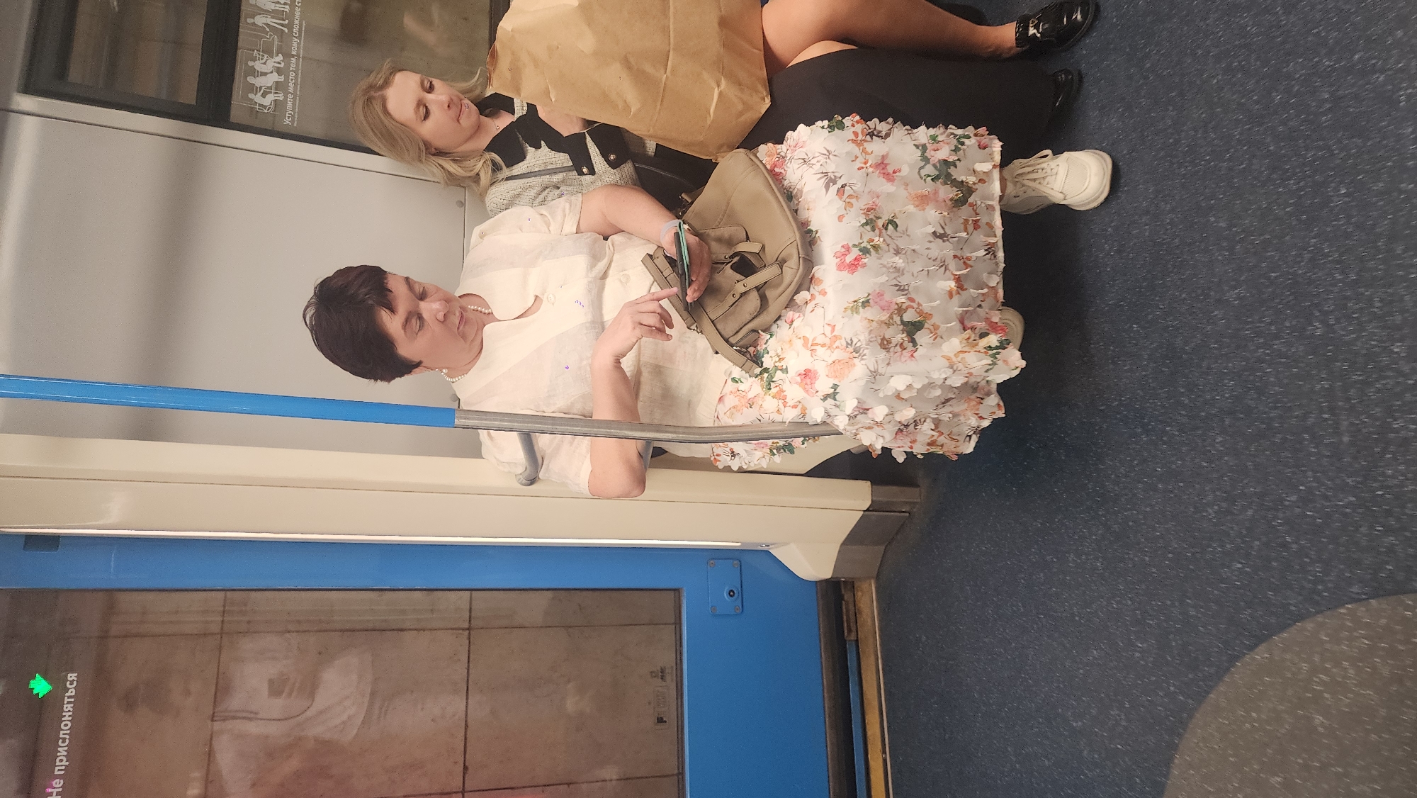 Two women are sitting next to each other on a train. The woman on the left has short dark hair and is wearing a light-colored blouse and pearl necklace, and is holding a smartphone in her hand. She has a beige handbag beside her, and is wearing a white floral skirt. The woman to her right is wearing a black jacket over a white and black top and is leaning against a large brown paper bag. She is wearing black shoes. The train car has a blue grab rail, a blue and white door, and a gray floor. A green arrow and Cyrillic text are visible on the door.