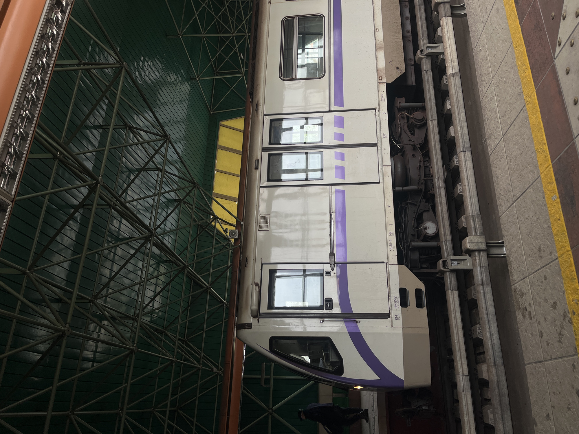 The image shows a subway train in the Sofia Metro, Bulgaria. The train is white with a purple stripe. The train is in a station. The platform is tiled. A yellow safety line is on the edge of the platform. The station has a green, structural roof. There is a dark shadow of a person.