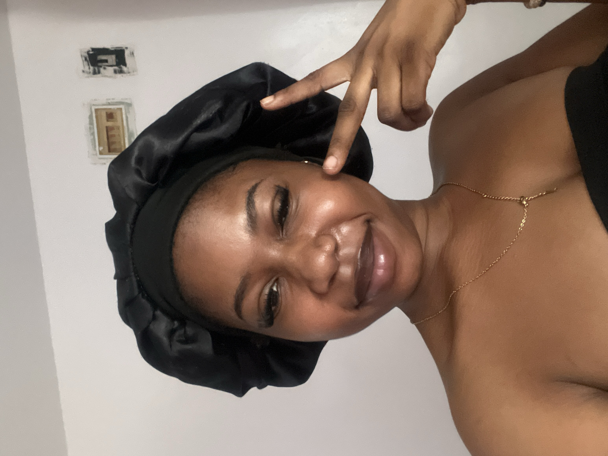 A young Black woman is posing for a selfie in what appears to be a bedroom. She is wearing a black satin bonnet on her head and a gold chain necklace. Her skin is smooth and appears to have a dewy glow. She is looking at the camera and smiling with her mouth slightly open. Her right hand is raised to her cheek with her index and middle fingers extended, forming a peace sign. The background is a plain white wall with a few framed items that are difficult to discern. There is a light source from the front, casting a soft glow on her face. The lighting suggests it is daytime. There is no information in the image to indicate the location as Jos, Nigeria.