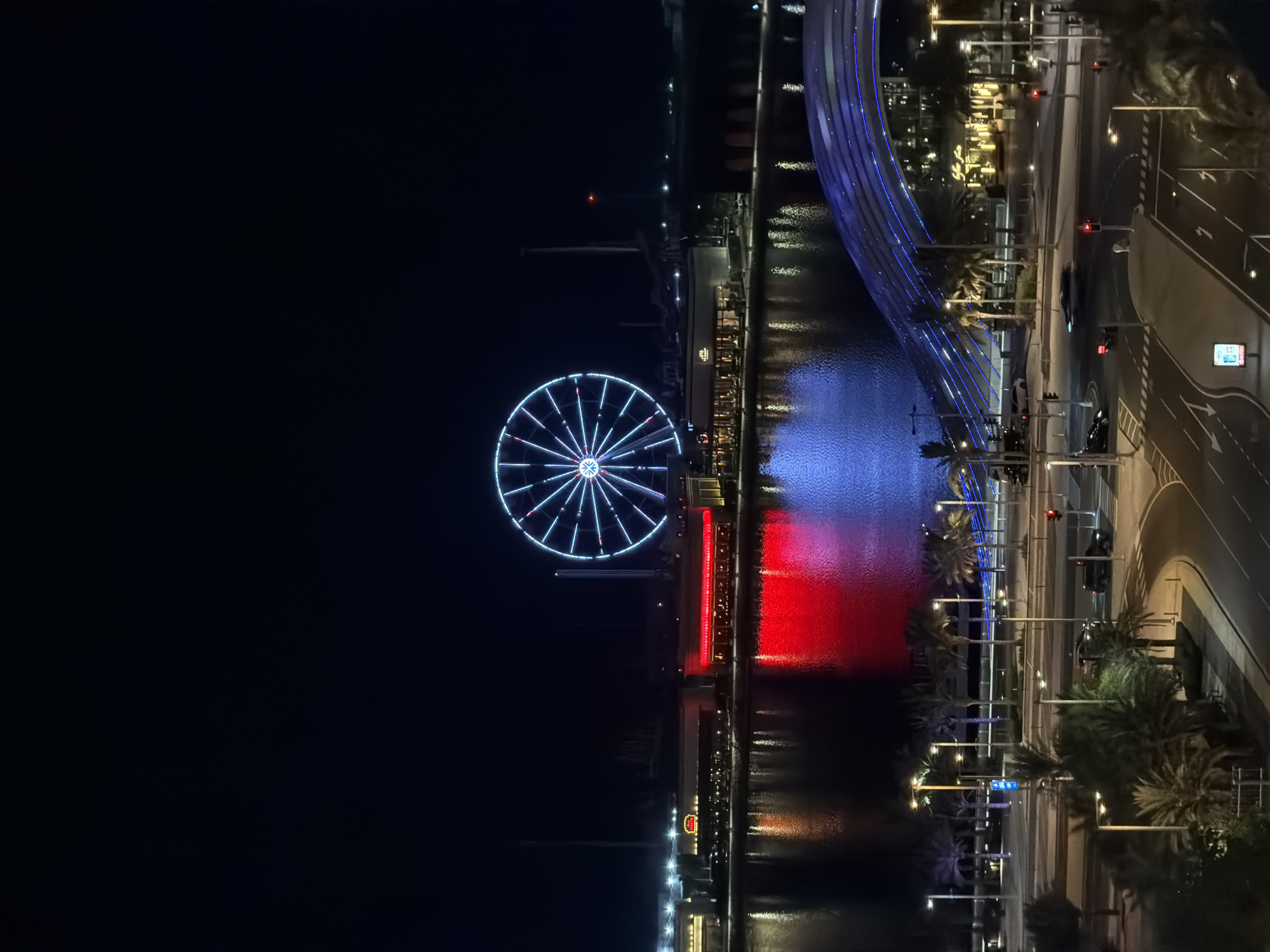 An aerial, long-shot, nighttime photograph shows a brightly lit Ferris wheel dominating the sky above a city skyline. The Ferris wheel is a large, circular structure with numerous spokes and is illuminated with blue and white lights. Below the Ferris wheel, a body of water reflects the lights of buildings and the Ferris wheel, with vibrant red and blue reflections visible on the water's surface. Buildings line the waterfront, some with glowing red and yellow lights, suggesting commercial establishments. In the foreground, a modern bridge with blue lights arches over a street. The street is visible with cars driving on it, and traffic lights are illuminated. Palm trees are scattered throughout the foreground and middle ground, adding to the urban landscape. The overall scene depicts a lively, modern city at night, characterized by bright lights and architectural features.