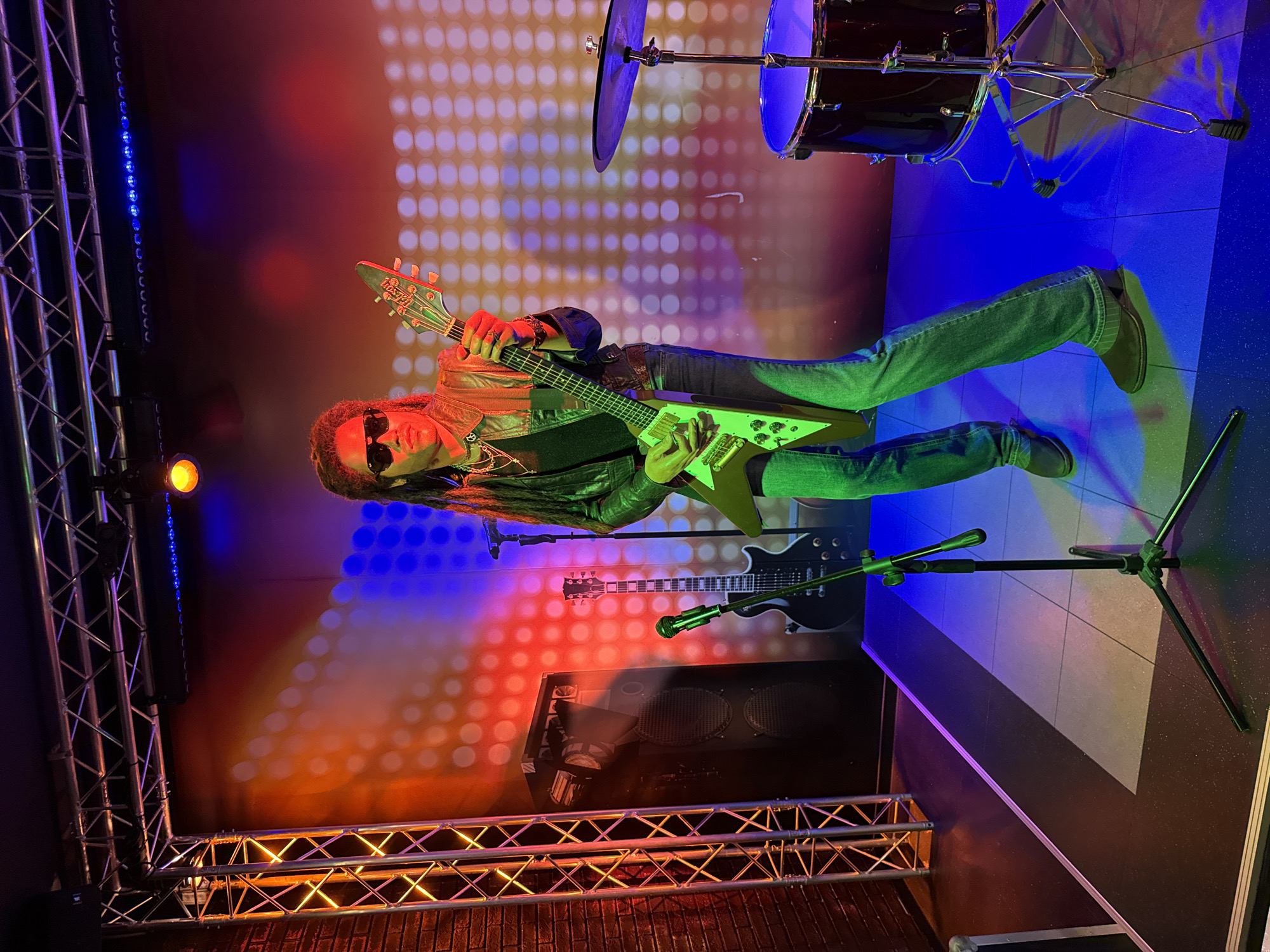 The image depicts a wax figure of a musician, resembling Lenny Kravitz, standing on a stage. The musician is wearing sunglasses, a leather jacket, and jeans, and is holding a V-shaped electric guitar. A black electric guitar, a microphone on a stand, and a drum set are also present on the stage. The backdrop features a gradient of red, orange, and blue lights with a pattern of illuminated circles. The stage is lit with colored lights, casting shadows and reflections. The scene appears to be an indoor exhibit, possibly a wax museum. There is no indication of the location being in Prague, Czechia.