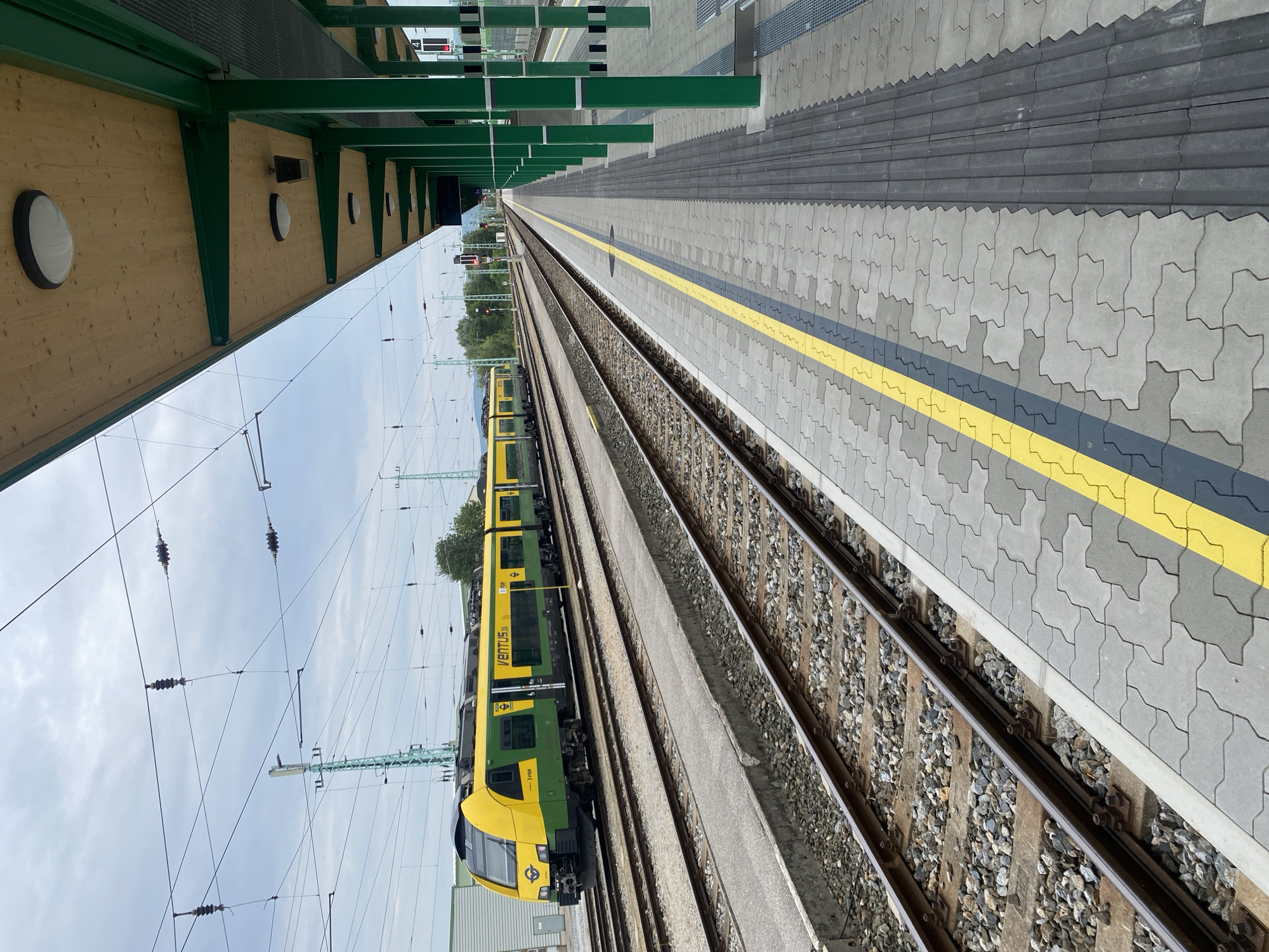 The image shows a train station platform in Wulkaprodersdorf/Vulkaprodrštof, Austria. On the left, a yellow and green train is stopped at the platform, adjacent to multiple train tracks. The train bears the inscription "VENTUS 35". The platform itself is paved with interlocking gray and yellow patterned tiles, with a tactile warning strip running along the edge closest to the tracks. To the right, a covered waiting area is supported by green pillars, with a wooden ceiling and overhead lighting fixtures. The sky is overcast with a few scattered clouds. Overhead power lines are visible across the scene, supported by tall green poles. The overall environment suggests a functional and modern train station.