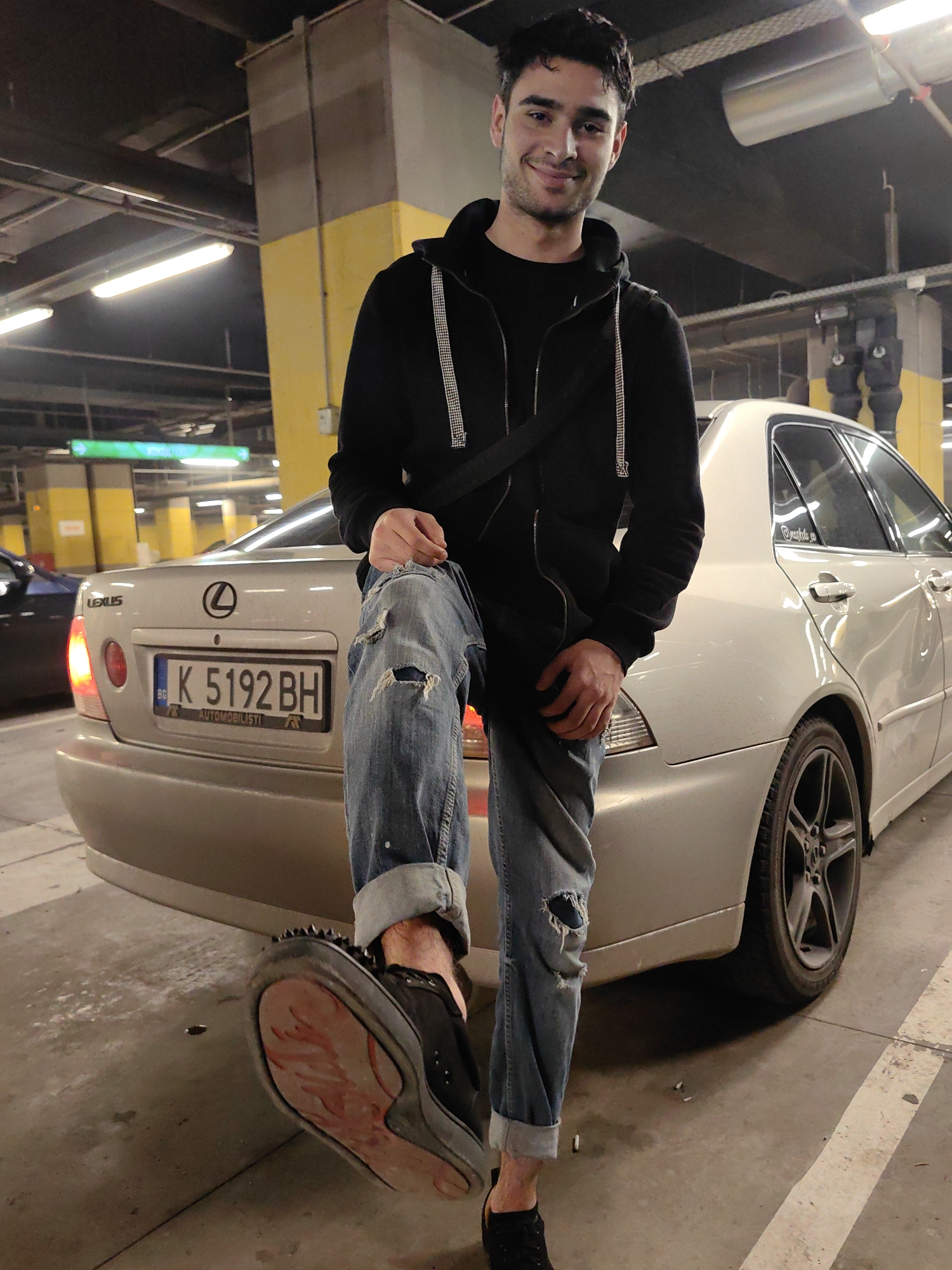 A young man poses in a parking garage in Sofia, Bulgaria, next to a silver Lexus sedan with license plate K 5192 BH. The man is wearing a black hooded sweatshirt, blue ripped jeans, and black studded shoes. He is smiling and looking at the camera, with one leg resting on the rear of the vehicle. The parking garage has exposed pipes and lighting fixtures on the ceiling, along with support beams painted yellow and grey.