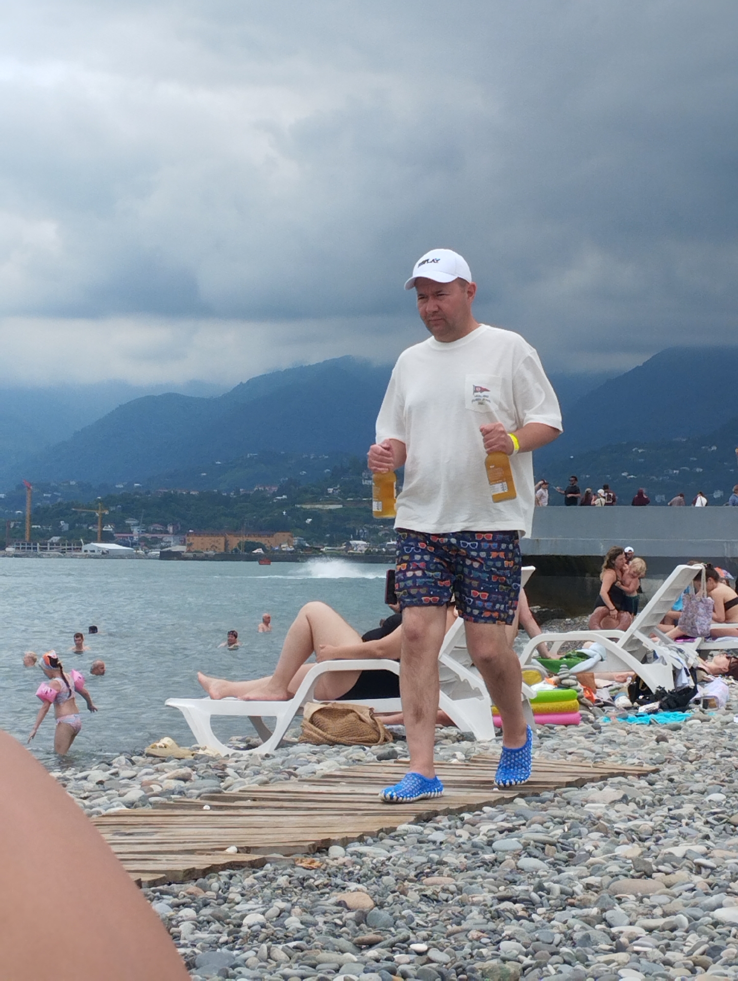 A man in a white t-shirt, shorts with a glasses pattern, and a white baseball cap walks along a pebble beach in Batumi, Georgia. He is holding two amber-colored bottles. In the background, people are swimming in the sea, and further back, mountains rise above a town. The sky is cloudy, suggesting overcast weather. Several white lounge chairs are set up on the beach, some occupied by sunbathers. A child in pink armbands is wading in the water. The scene appears to be a casual day at the beach, possibly in the afternoon, with a relaxed atmosphere. The man's expression is neutral as he walks. Visible text on the man's cap appears to say "REPLAY".