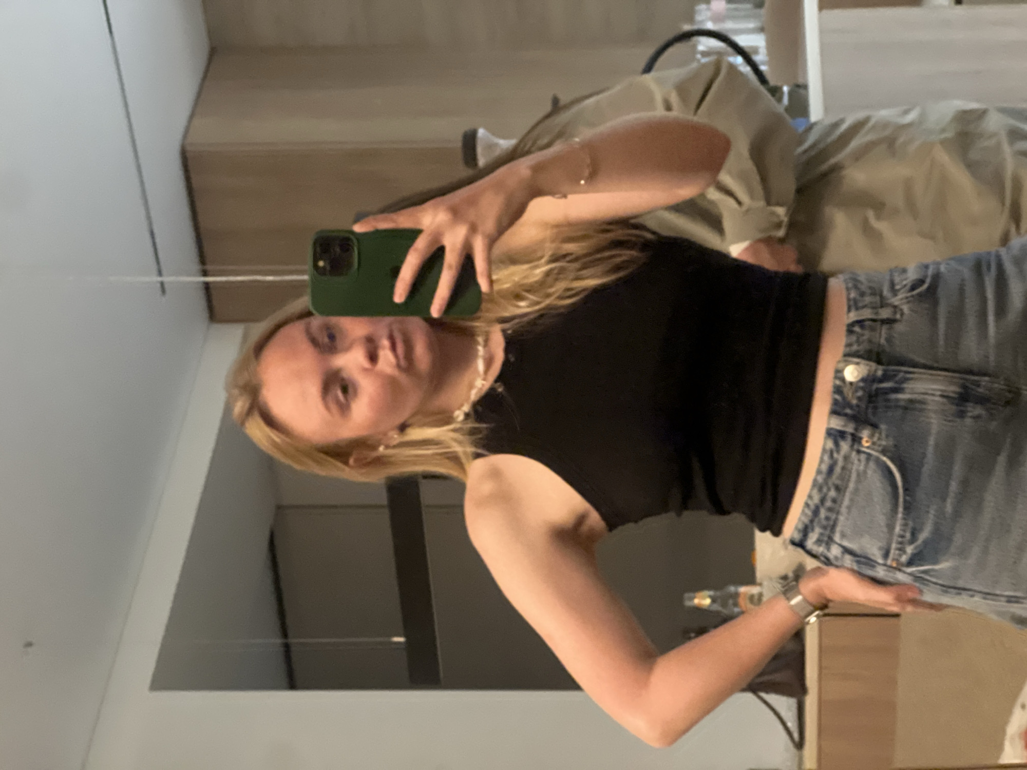 A young woman with blonde hair takes a mirror selfie. She is wearing a black top, blue denim shorts, and a pearl necklace. She holds a green phone up to the mirror with her right hand. Her left hand is near her waist. The background shows part of a room with furniture and walls. The location is unknown and there is no indication this image was taken in the city of Рубцово, Russia.