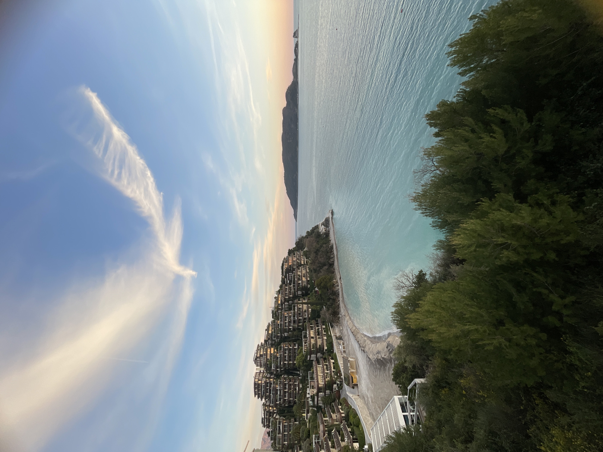 The image displays an elevated panoramic view of a coastal scene in Budva, Montenegro, specifically overlooking the area towards Sveti Nikola Island. The sky, a clear light blue, features elongated, wispy white clouds and a faint contrail. The horizon transitions from blue to soft orange and pink, indicative of either sunrise or sunset.
To the left, a prominent hillside descends towards the sea, densely populated with a terraced complex of modern, multi-story buildings, consistent with the architecture of Dukley Gardens. These buildings are integrated with abundant green vegetation and trees. Below the hillside structures, a light-colored, crescent-shaped beach stretches along the coastline, with white foam visible where the gentle waves meet the sand.
The Adriatic Sea dominates the midground and right, exhibiting clear, light turquoise water near the shore that gradually deepens to a bluer hue further out. Subtle ripples are visible on the water's surface. A small, indistinct red object, possibly a buoy, floats in the distant water to the right.
On the distant horizon to the right, a large, dark, elongated landmass, identified as Sveti Nikola Island, is silhouetted against the lighter sky, with smaller rock formations visible beyond its eastern tip. The immediate foreground of the image is filled with dense, dark green foliage, primarily coniferous trees, indicating the elevated vantage point from which the photograph was taken. No individuals, specific actions, or interactions are discernible within the scene.
