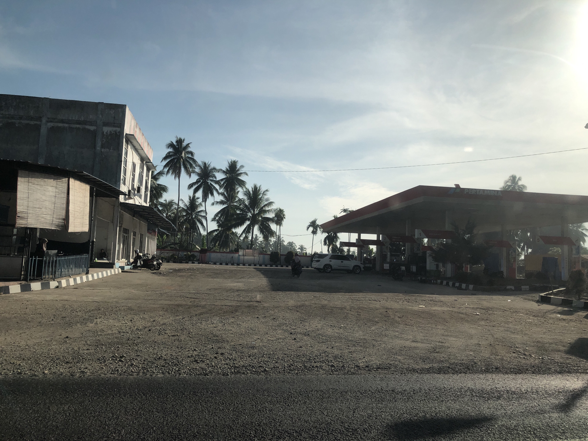 This image captures an outdoor scene in Kutalawah, Indonesia, featuring a Pertamina gas station and an adjacent multi-story building under a bright, partly cloudy sky. The time of day appears to be late morning or early afternoon, given the bright sunlight and visible shadows.
On the left side of the frame, there is a concrete building, possibly unfinished or unpainted, with a lower section featuring windows and an attached structure with a bamboo-like blind. Near this building, several motorbikes are parked, and at least one person is visible behind a metal fence. Another individual on a motorbike is seen driving or waiting in the mid-ground.
The right side of the image is dominated by the Pertamina gas station, identifiable by the "PERTAMINA" logo prominently displayed on its red-roofed canopy. Underneath the canopy, several gas pumps are visible, and a white SUV is parked. Numerous tall palm trees are scattered throughout the scene, especially behind the gas station and the main building, contributing to a tropical setting. The ground consists of a rough, gravelly surface in the foreground and around the station, with sections of darker asphalt. Black and white striped road barriers are visible around the edges of the paved areas. The overall impression is that of a typical roadside stop or service area in a tropical region.
