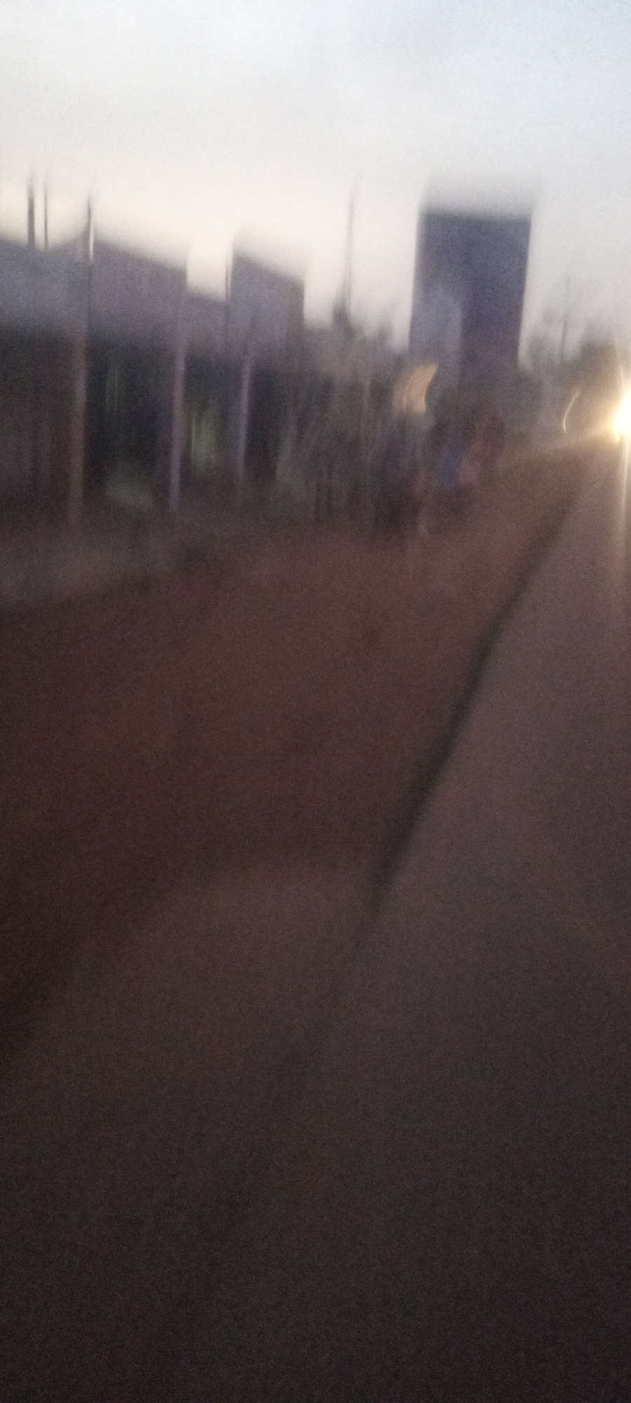 The image depicts a blurred view of a street in Kabala, Nigeria, likely taken at dusk or dawn. There are buildings on the left side of the street, appearing somewhat indistinct due to the blur. A light source, possibly a vehicle's headlight, is visible in the distance on the right side. The road itself occupies the foreground and midground, appearing dark and textured. There may be people present near the buildings, but they are not clearly discernible.