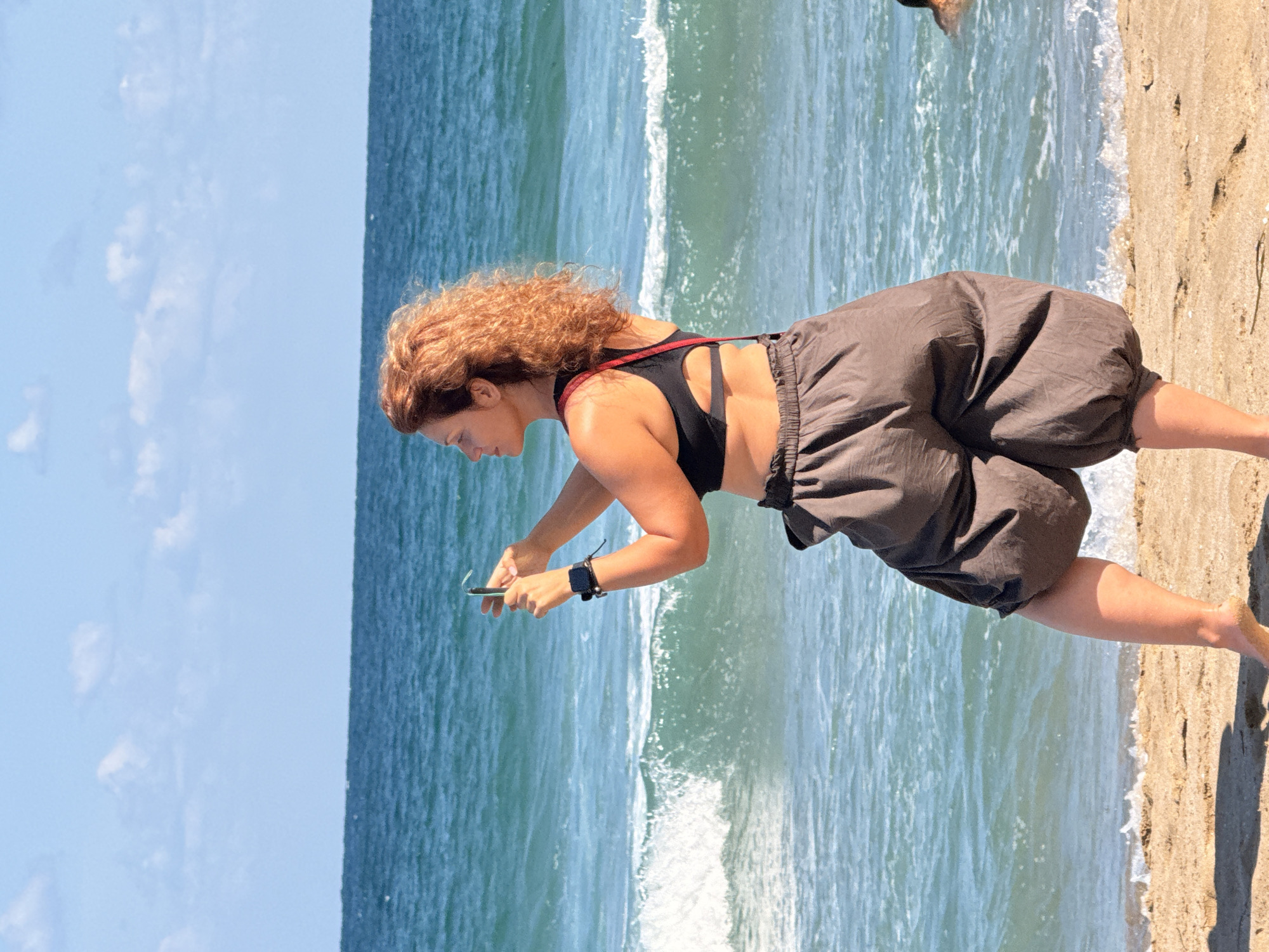 A woman with curly reddish-brown hair is standing on a sandy beach, facing left, holding a smartphone in her hands as if taking a picture. She is wearing a black sports bra and loose-fitting, dark brown, voluminous shorts. A red strap is visible over her left shoulder. Her left arm is bent at the elbow, with her hand holding the phone. She has a smartwatch on her left wrist. The ocean is behind her, with blue waves breaking and white foam on the shore. The sky is a clear blue with scattered white clouds. The scene appears to be a sunny day at the beach. The location is the city of Sozopol, Bulgaria.
