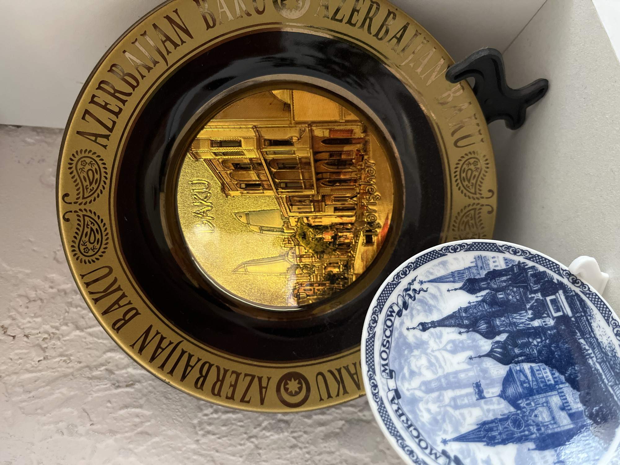 The image shows two decorative plates displayed on a white surface. The larger plate, positioned behind and to the right, is gold with a dark brown rim. It features text around the edge that reads "AZERBAIJAN BAKU" and "BAKU AZERBAIJAN" in an ornate font, interspersed with paisley designs. The center of this plate depicts a golden cityscape with prominent modern architecture, including a tall, flame-like skyscraper, and older, traditional buildings. Below the cityscape, the word "Baku" is visible. This plate is propped up by a small black plastic stand.
In the foreground and to the left is a smaller, white plate with blue and white decorative patterns. The central image on this plate is a detailed blue and white illustration of a city skyline, featuring domed buildings and towers that strongly resemble the architecture of the Moscow Kremlin, specifically St. Basil's Cathedral. The word "MOSCOW" is written in blue script across the top of this plate, with decorative banners on either side.
The background is a textured white wall. There are no people or animals visible in the image. The plates are decorative souvenir items. The image does not contain any information related to the city of Krasnyye Barrikady, Russia.