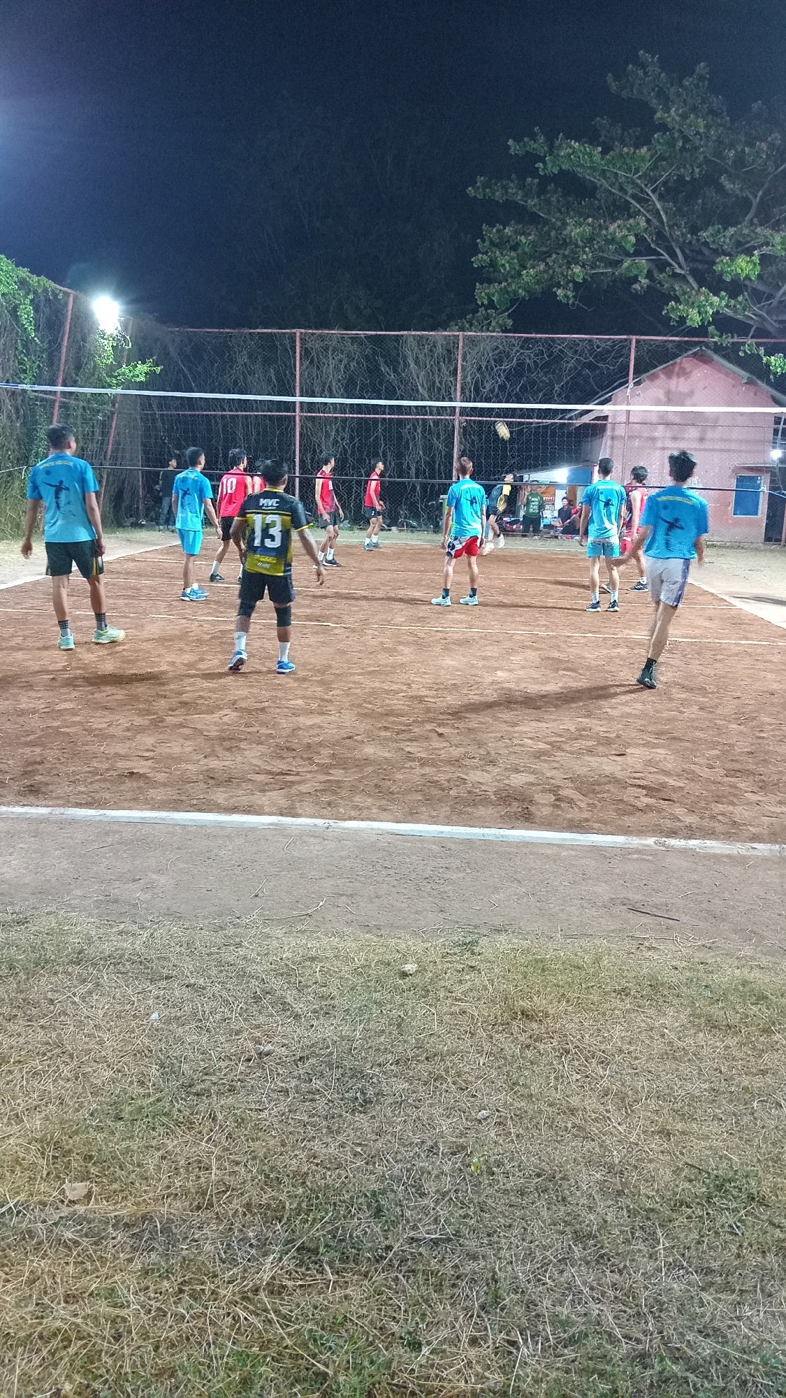 The image depicts a night-time outdoor volleyball game taking place on a dirt court in Prambangan, Indonesia. The court is illuminated by artificial lighting, casting shadows across the sand-colored surface. A net divides the court, with players on both sides engaged in the game.
There are at least ten visible individuals, mostly young men, dressed in athletic attire. Several players are wearing blue shirts, while others are in red or yellow and black. One player in the foreground is wearing a yellow and black jersey with the number "13" clearly visible on the back.
The environment surrounding the court includes a chain-link fence bordering the playing area, with trees and foliage visible beyond it, suggesting a rural or semi-rural setting. In the background, a building with a pinkish hue and a red roof can be seen, also illuminated by artificial light. The sky is dark, indicating it is nighttime. The grass area in the foreground appears somewhat dry and unkempt. The overall scene suggests a casual or amateur sporting event.