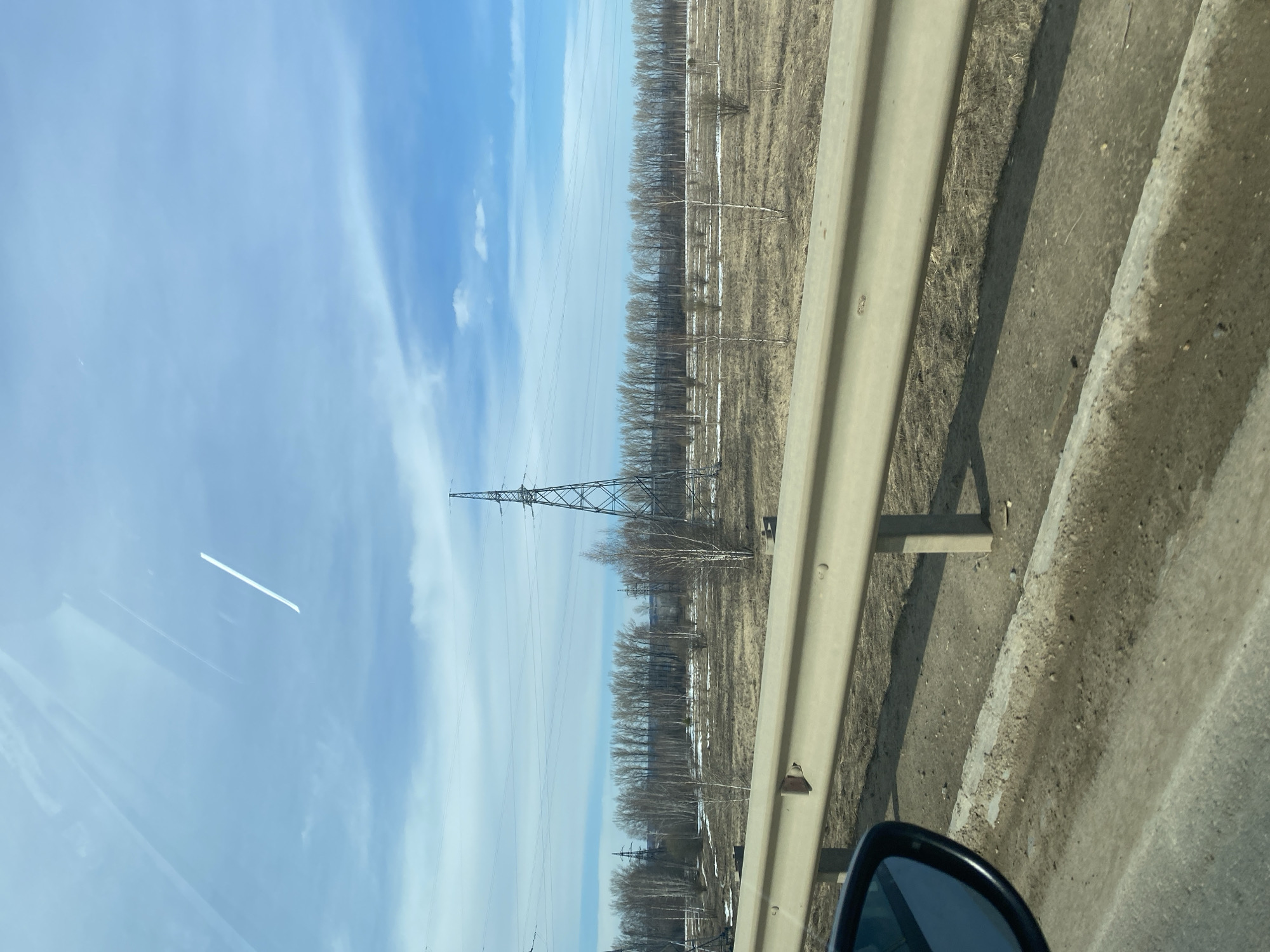 The image depicts a scene from a moving vehicle, potentially on a road adjacent to a field or open area. A guardrail runs along the right side of the frame. Beyond the guardrail, the landscape is characterized by sparse vegetation, potentially early spring or late autumn conditions. A power line tower is visible in the distance, stretching towards a blue sky with light clouds. The vehicle's side mirror is visible in the lower right corner of the frame. The location is speculatively Kueda, Russia.