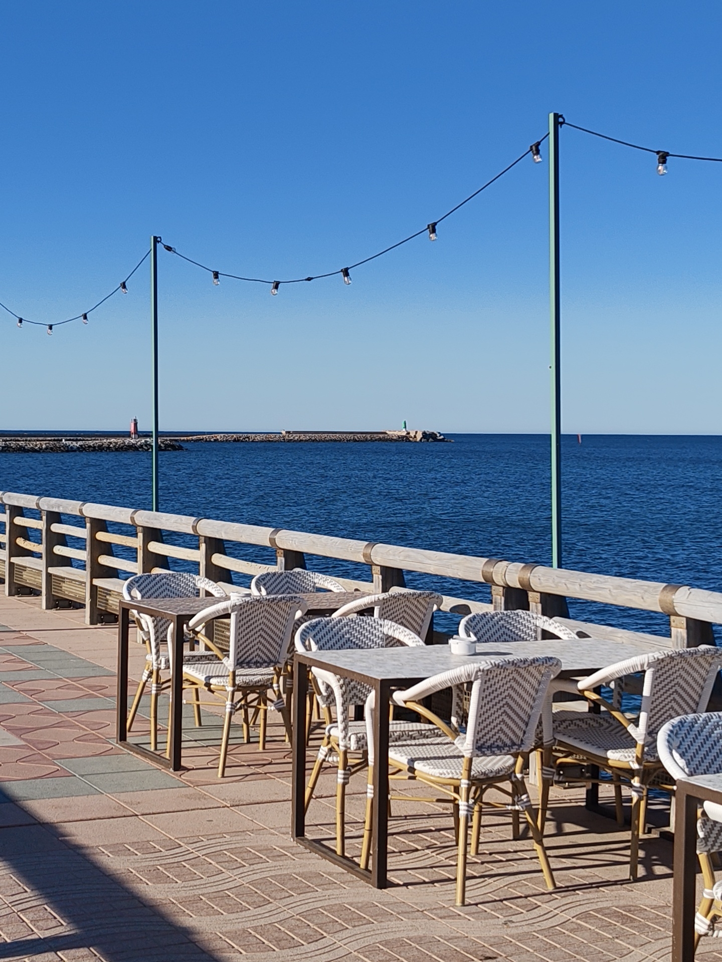 This image captures a peaceful, sunny outdoor dining area by the sea, likely in Dénia, Spain. The scene is set on a paved promenade overlooking clear blue water, with no people visible, conveying a sense of calm and readiness for guests.
In the foreground, several outdoor tables with dark metal frames and light-colored tops are arranged, accompanied by woven chairs featuring white and light grey patterns on their seats and backs, supported by light brown, possibly bamboo-style, legs. On one table, a small white ashtray is visible. The ground is paved with multi-toned tiles in patterns of brown, grey, and reddish hues, displaying elongated shadows from the tables and chairs, indicating bright daylight.
A sturdy wooden railing with horizontal beams and metal accents separates the dining area from the tranquil, deep blue sea. Across the clear azure sky, string lights with exposed bulbs are strung between two tall, thin green poles, suggesting the space is also used during the evening.
In the mid-ground, a stone breakwater or pier extends into the sea, featuring two small lighthouses: a red one positioned on the left and a smaller green and white one further right. The horizon line is clear, and the water appears calm with gentle ripples, reflecting the bright conditions. The overall atmosphere is inviting and serene, typical of a coastal resort or restaurant awaiting its patrons. No text is visible in the image.