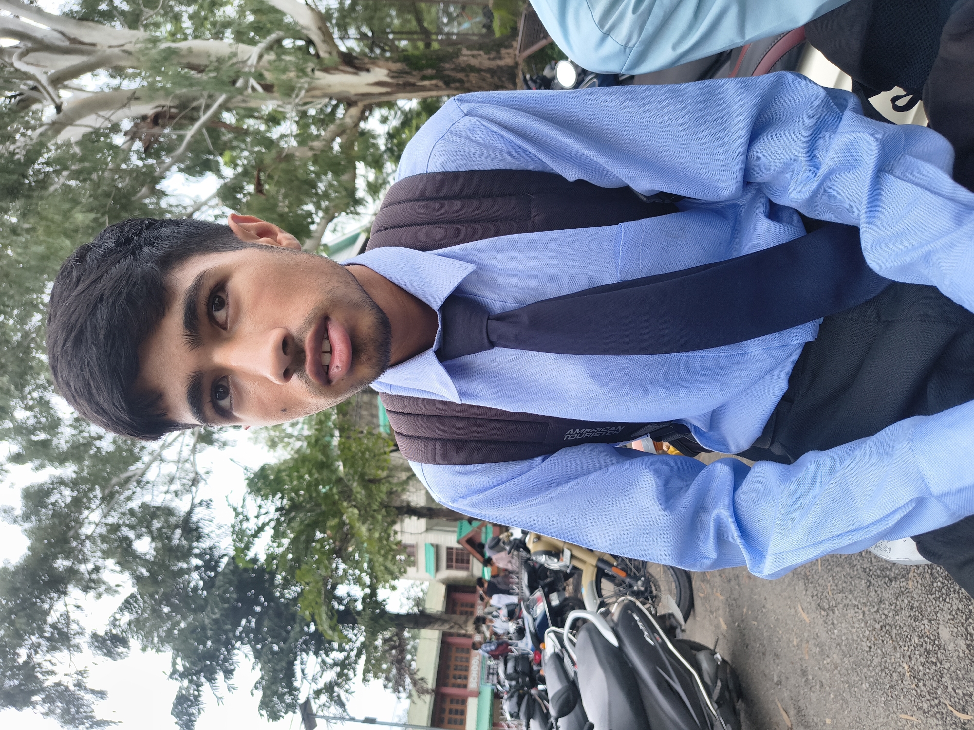 A young South Asian male, appearing to be in his late teens or early twenties, is captured in a medium close-up shot outdoors. He has dark hair, dark eyes, and a light complexion, with a faint stubble on his face. He is wearing a light blue collared shirt, a dark tie, and a dark backpack. His mouth is slightly open, and his gaze is directed slightly to his right.
The background suggests an urban environment with a large tree with white bark and leafy branches behind him. In the middle ground, there are buildings with visible windows and signage, including what appears to be a "SCHOOL" sign. A significant number of parked motorcycles and scooters are visible in the foreground and mid-ground, indicating a location where transportation is common. The ground surface is a mix of gravel and asphalt.
The overall impression is of a student, possibly leaving or arriving at a school in Dharamshala, India, given the presence of the school building and the common use of two-wheelers for transport in such locations. The lighting suggests it is daytime, possibly overcast.