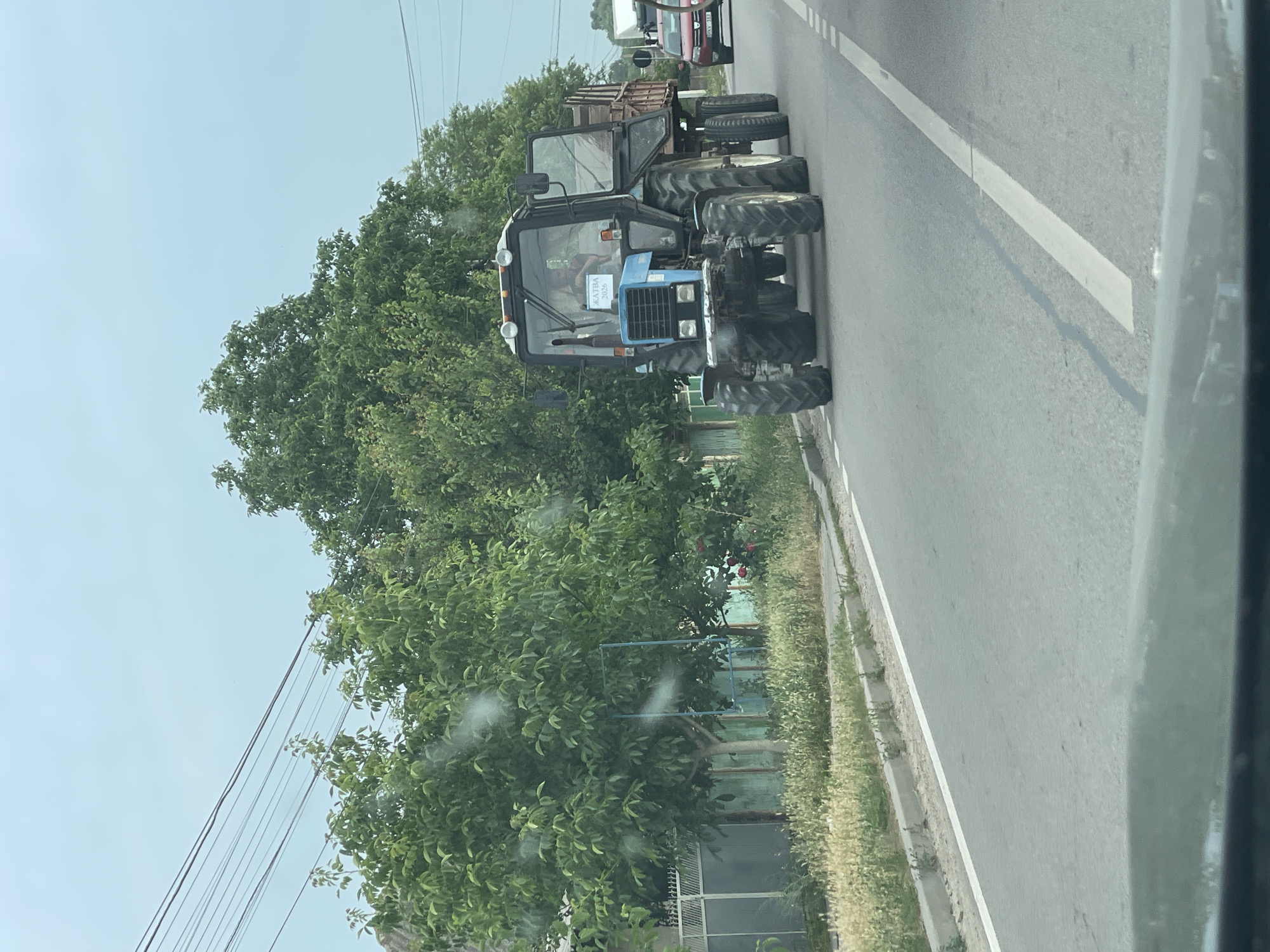 The media file depicts a blue tractor on a road in Congaz, Moldova. The tractor is the primary subject, with its large tires and distinctive blue cab being prominent. Inside the cab, a person can be seen, though their features are not clear. A sign is visible on the front of the tractor with text that reads "ЖАТВА 2020," indicating that it is participating in or related to the 2020 harvest.
The setting appears to be a rural or semi-rural area, with trees and vegetation lining the road. Power lines are also visible in the background, suggesting infrastructure present in the area. The road itself is paved with a white line marking its edge. In the distance, other vehicles, including what appears to be a truck, are present, indicating this is an active road.
The lighting suggests it is daytime, with a bright sky, though the overall impression is somewhat overcast. The image is taken from a moving vehicle, as indicated by the slight blur and the perspective. There are no strong emotional cues conveyed by the image itself, beyond the practical depiction of a working tractor in its environment. The focus is on the agricultural machinery and its immediate surroundings in the context of a harvest event.